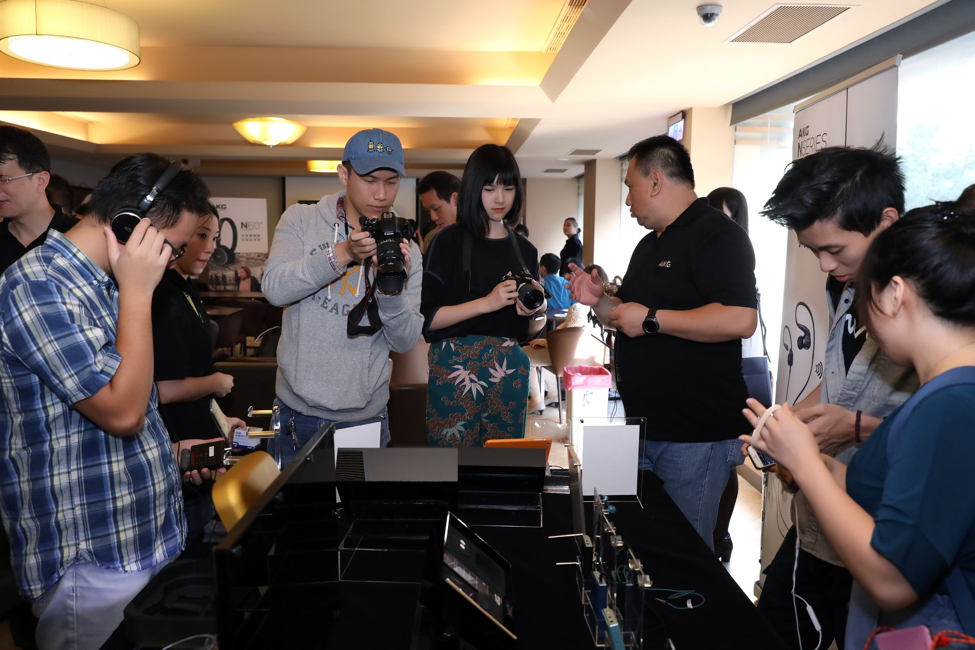 People test electronic devices at an event. Some wear headphones, others hold smartphones. Indoors, bright lighting.