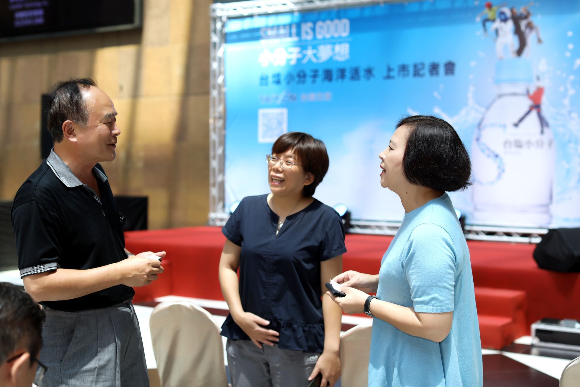 Three people conversing in front of a banner; one holds a water bottle.