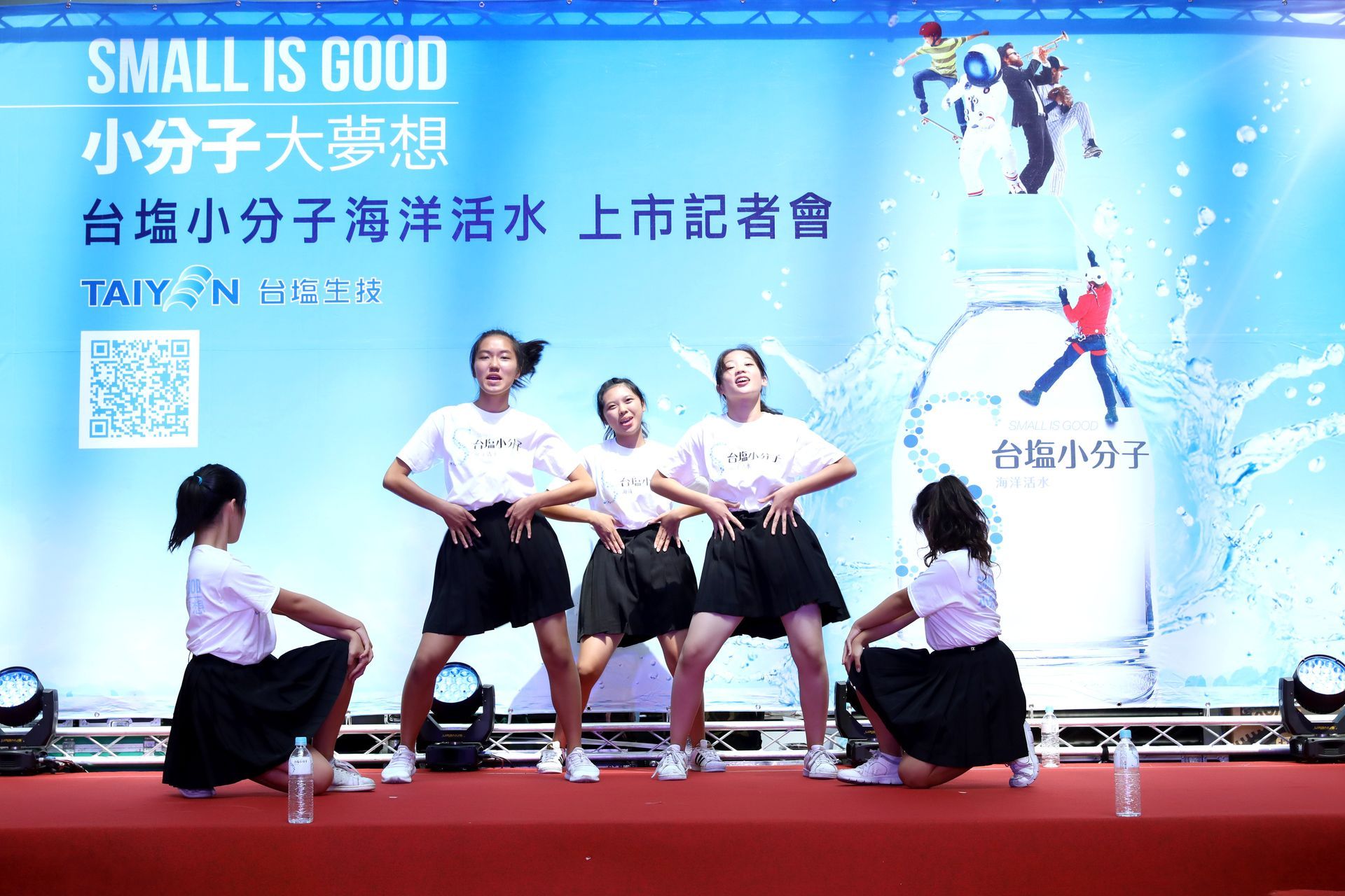 Dancers in white shirts and black skirts perform on a stage with a blue backdrop that promotes a water brand.