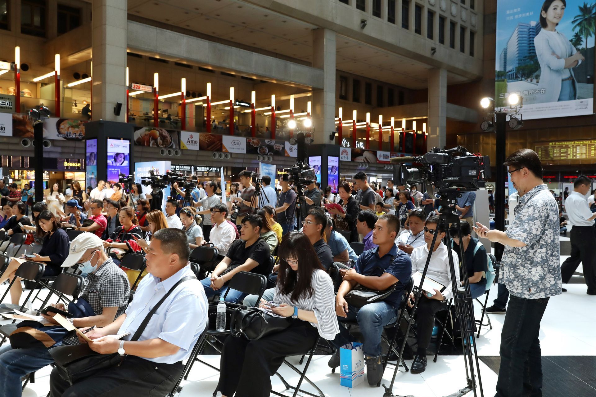 Large crowd seated and standing, many with cameras, attending an event in a large indoor space.
