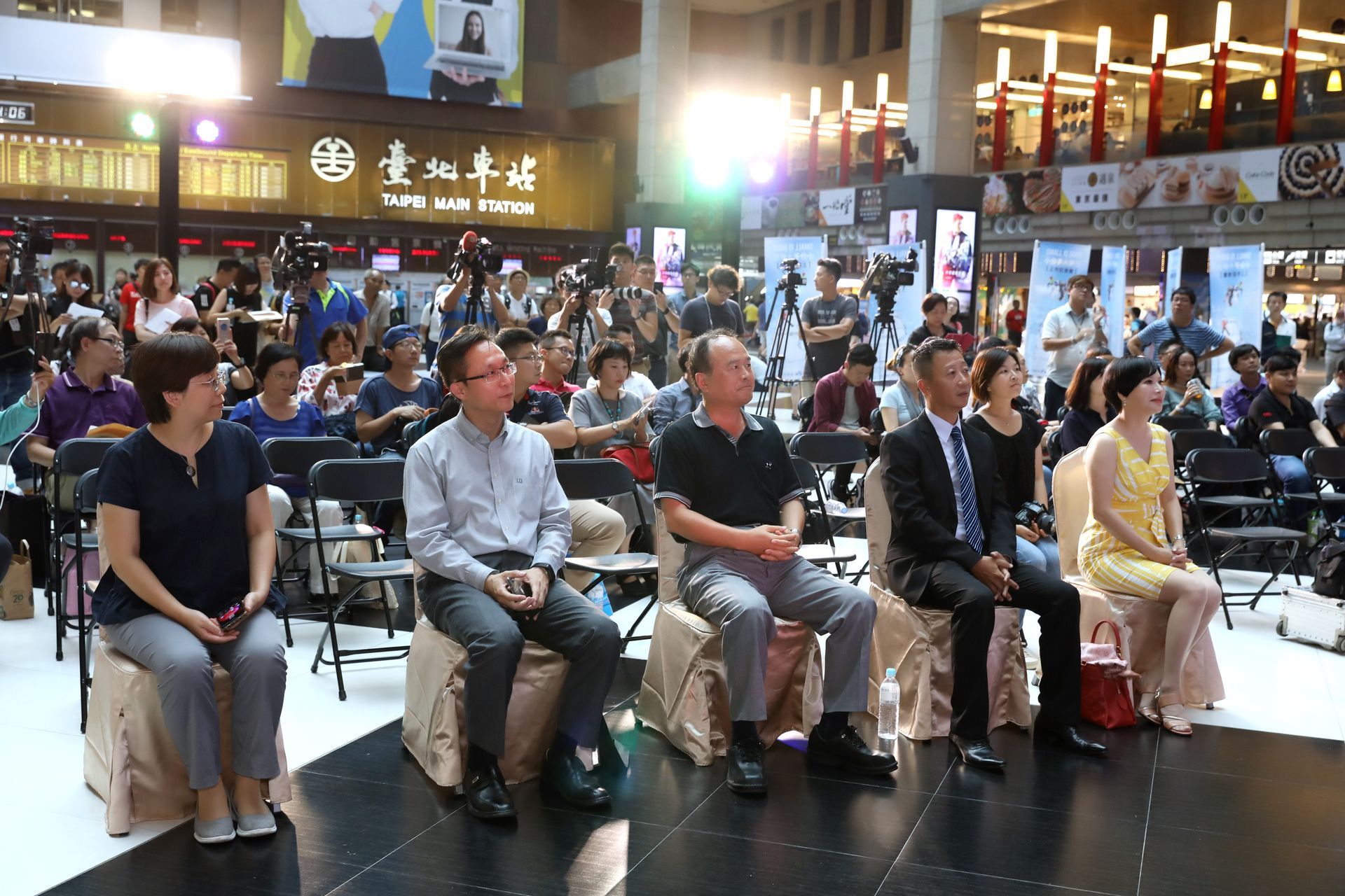 People seated at an event, with audience and cameras in background, in a large open space.