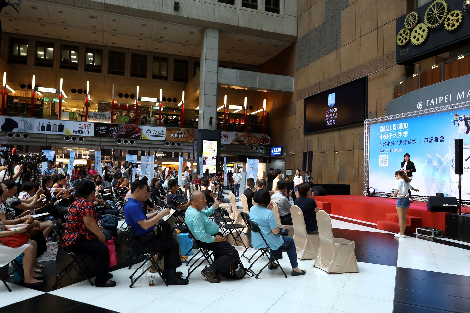 A presentation on a stage, audience seated. The setting appears to be a large public space, with a screen showing information.