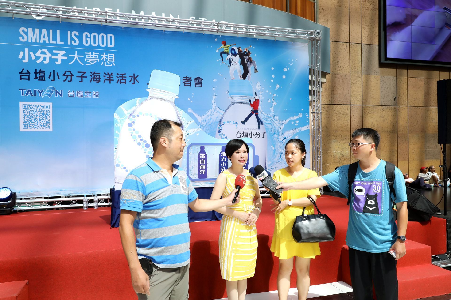 People on stage, promoting bottled water. Backdrop with text, blue, white, and red. Two people hold microphones.