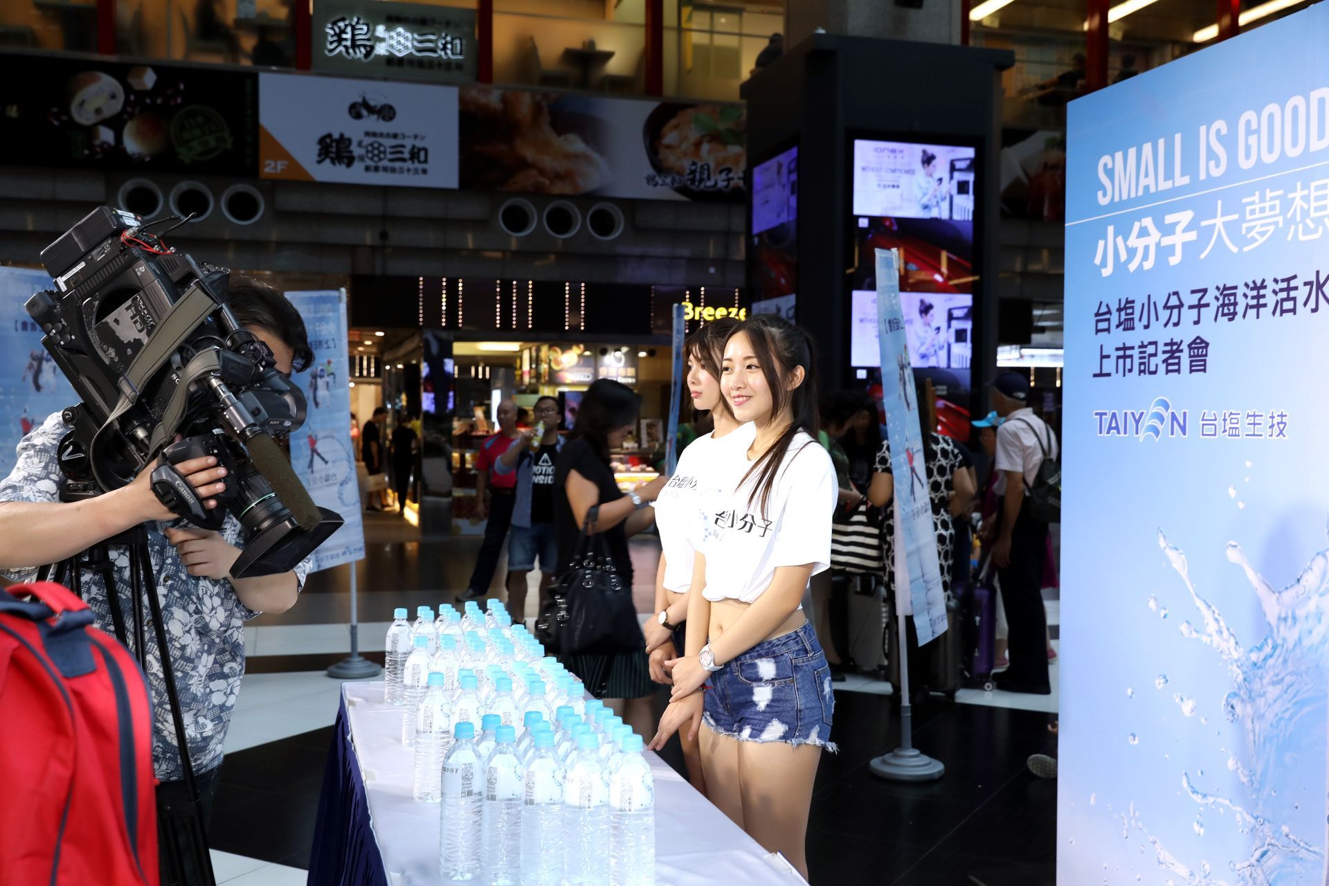 A videographer films two women posing next to a display of bottled water in a public space.