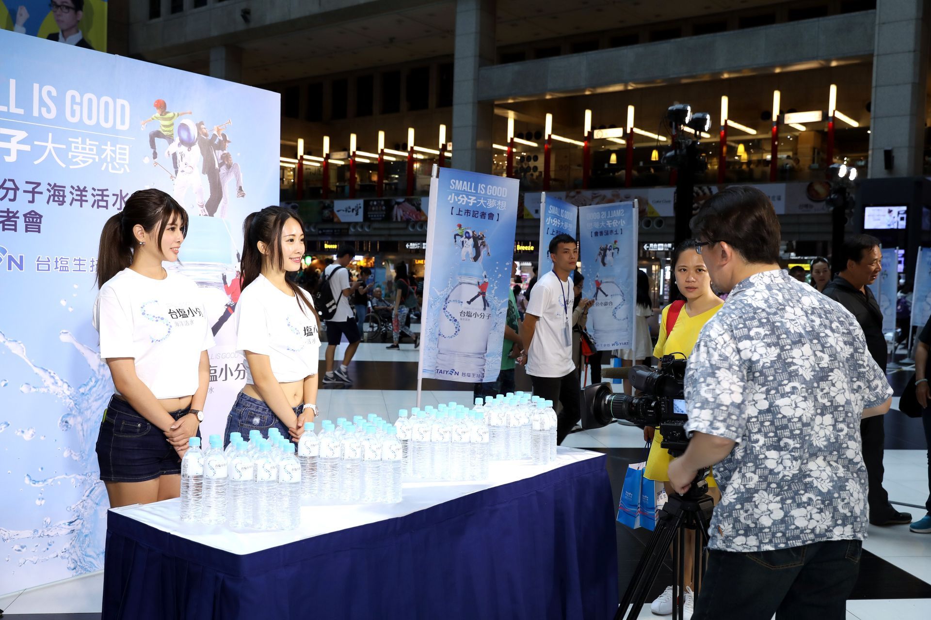 Two women pose at an event table with water bottles. A man films them. Event backdrop: 