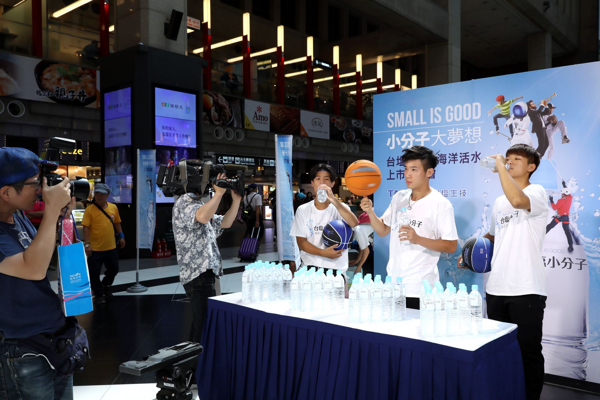 People at an event with a table of water bottles and a basketball, being filmed by cameras.
