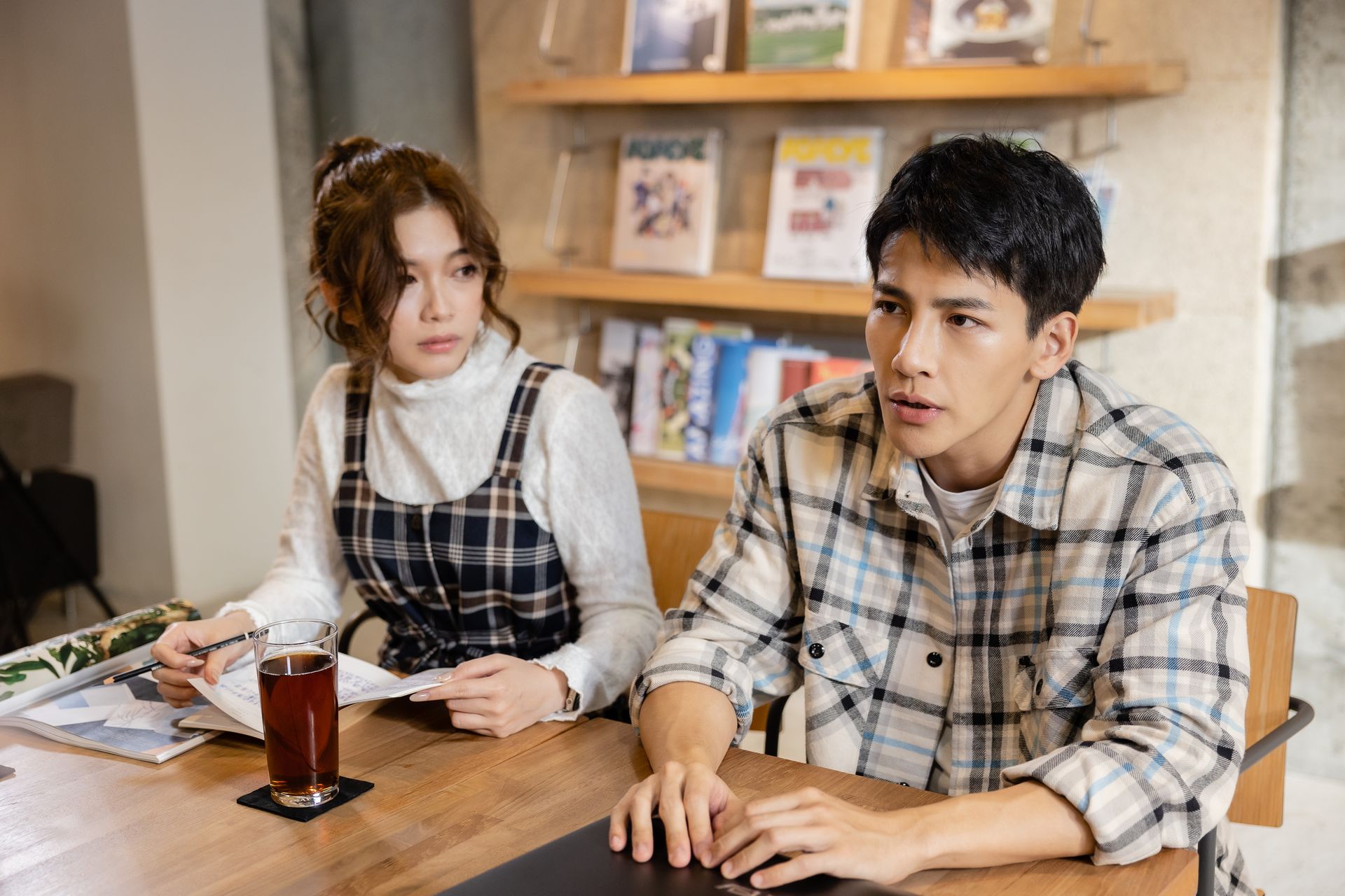 Woman and man at a table in a cafe, looking concerned. Woman in plaid jumper, man in plaid shirt.