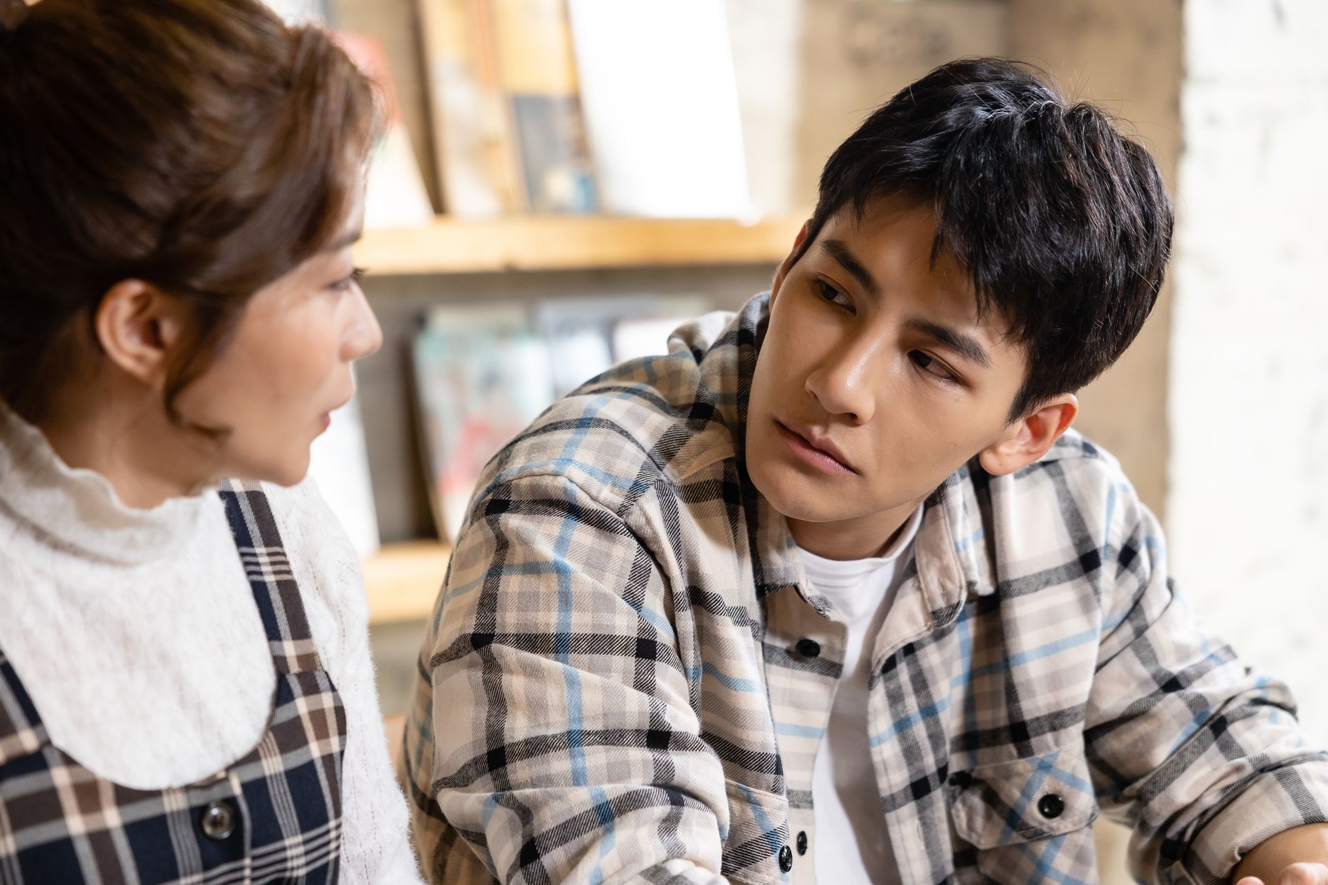 Woman in a patterned dress speaks to a man wearing a plaid shirt. They are indoors, possibly a cafe.