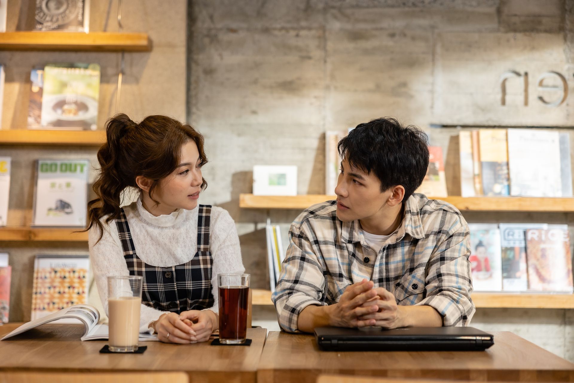 A couple at a table in a cafe, looking at each other. Bookshelves in the background; drinks on the table.