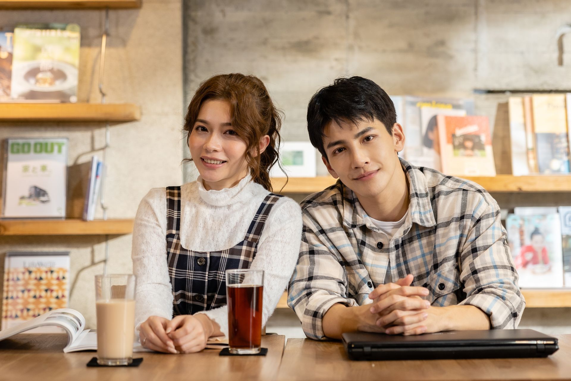 Woman and man smiling at a table with drinks and a laptop, in a cafe with bookshelves.