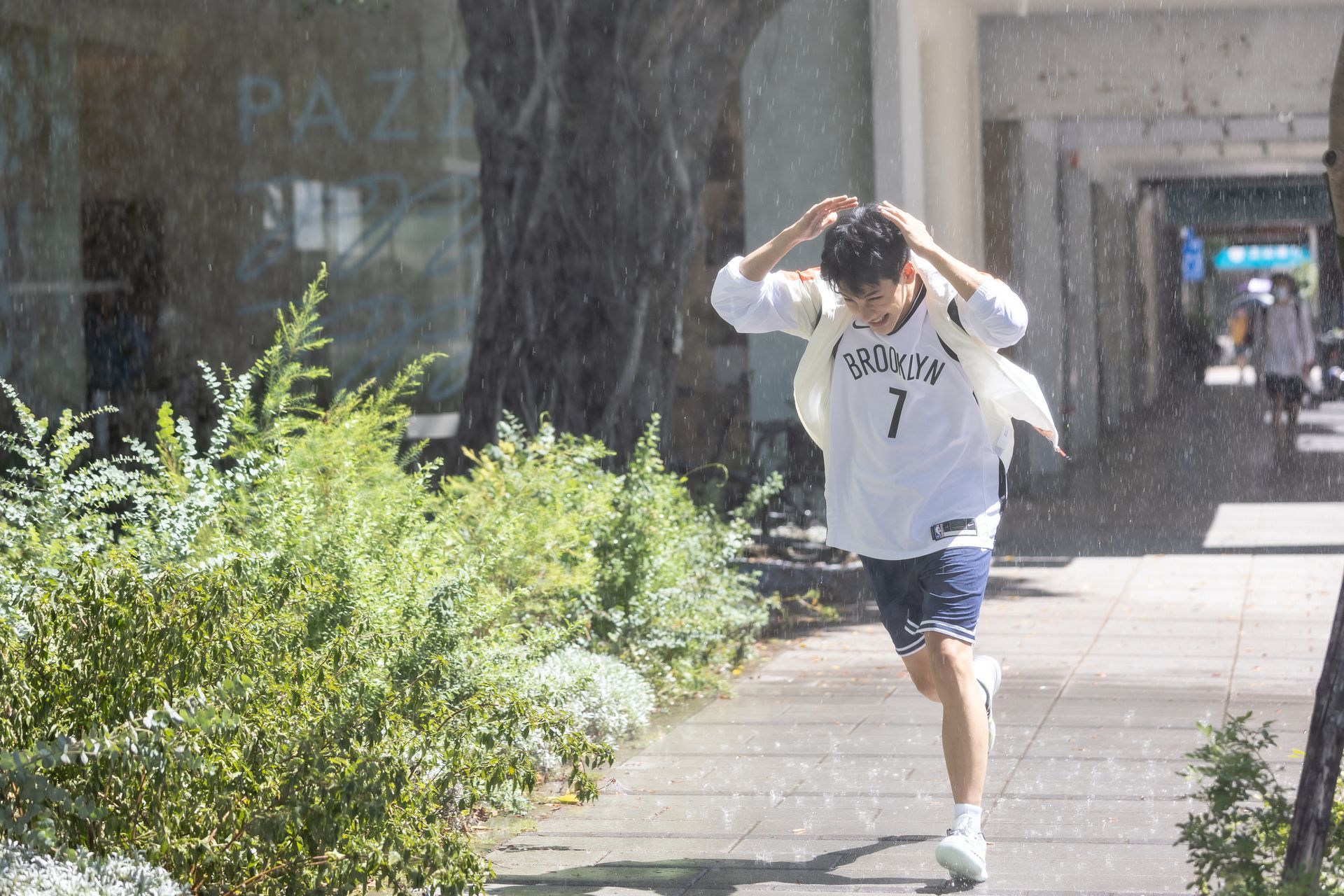 Person running in the rain, covering their head. Sidewalk next to green bushes and a building.