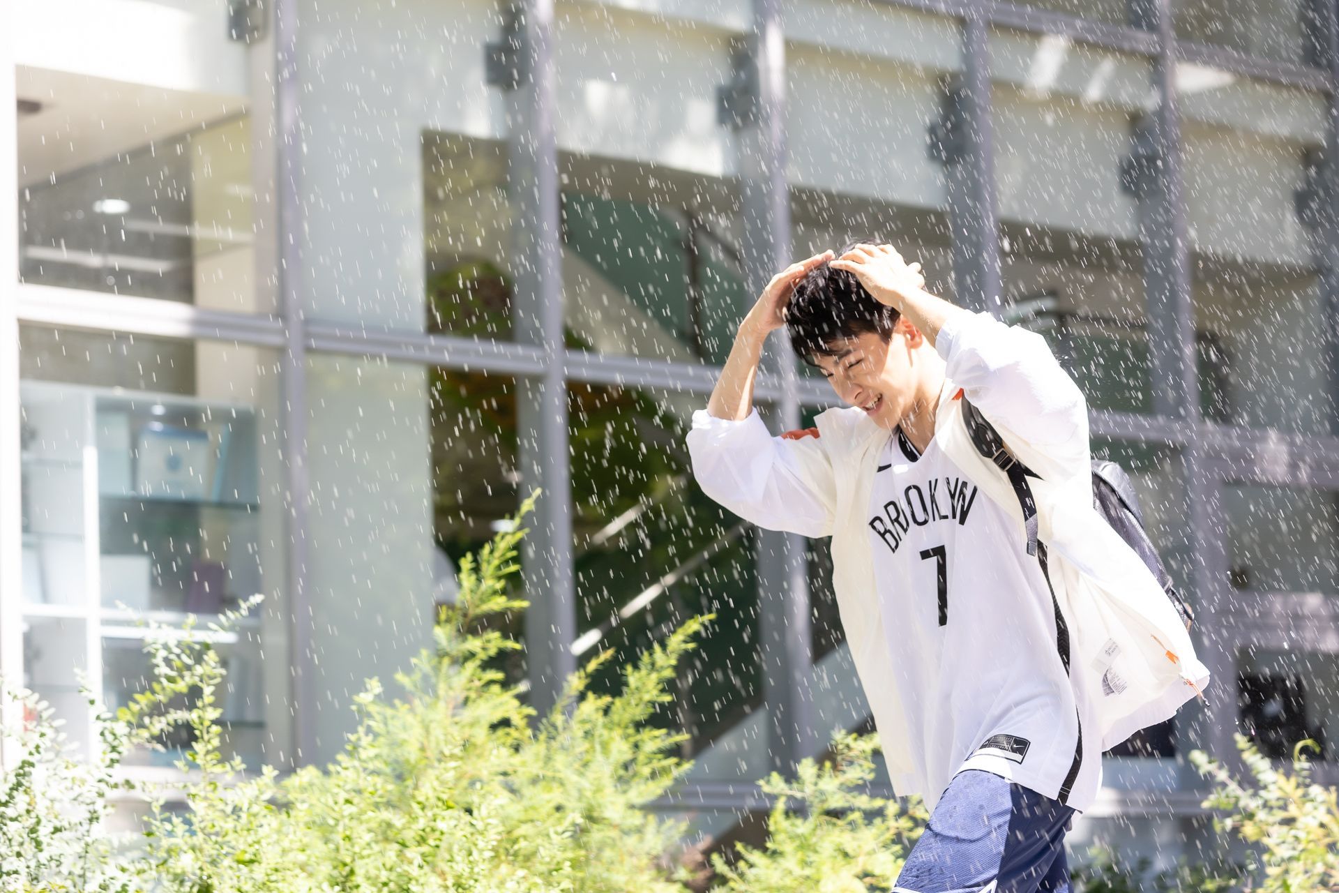 Person outside in the rain, wearing a basketball jersey, shielding head with hands, near a building.