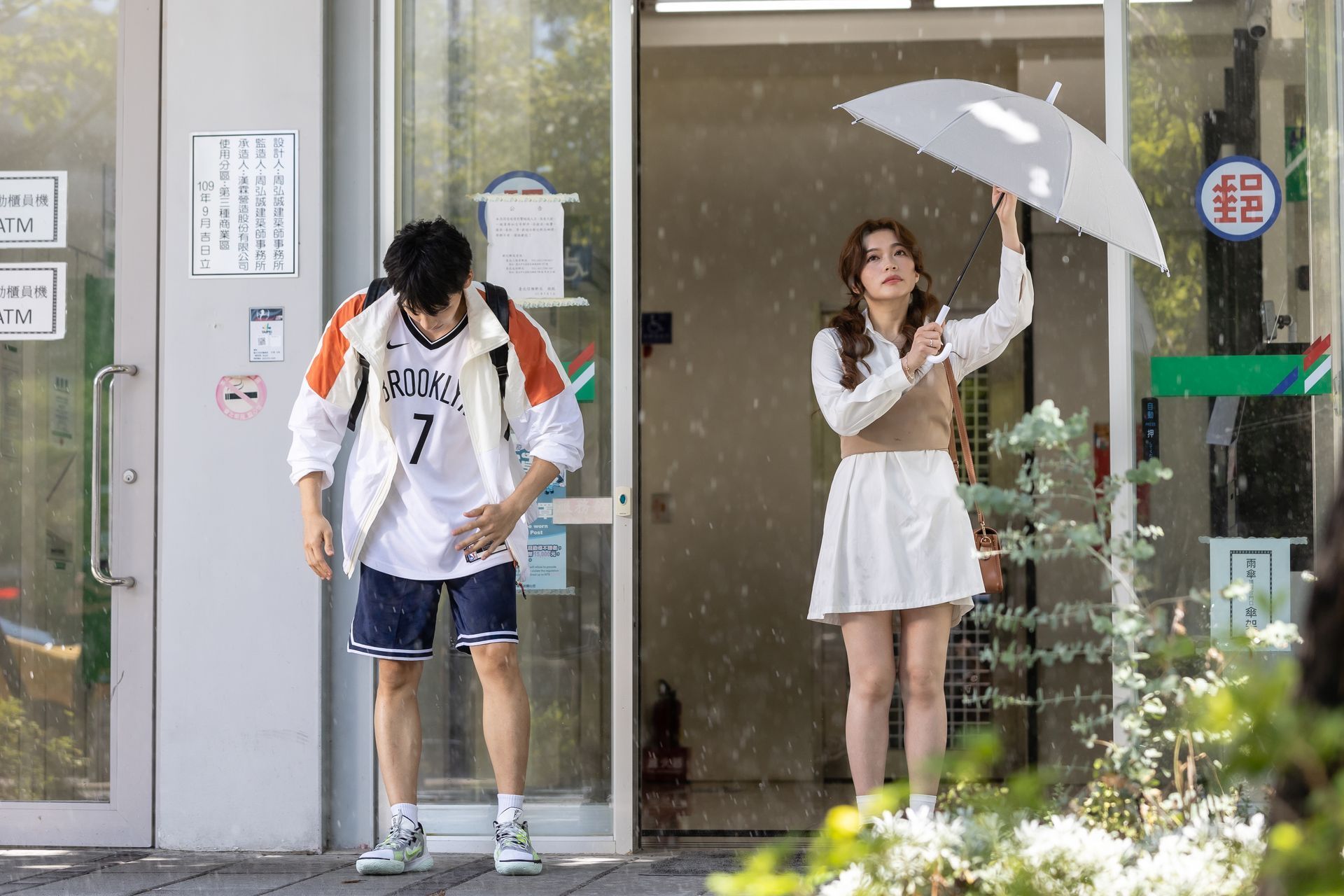 Man in sportswear hunches over, while woman holds umbrella; both outside a store in the rain.