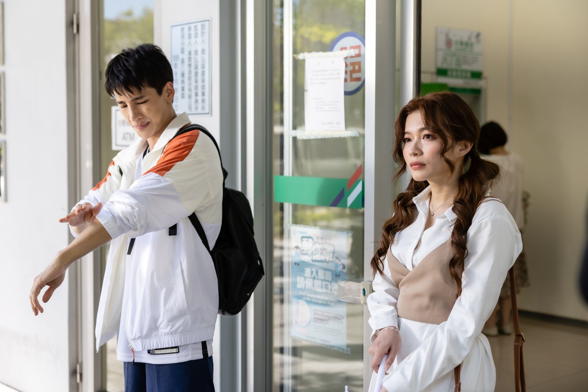 Man adjusting sleeve; woman near doorway. Both looking off-camera. Bright setting.