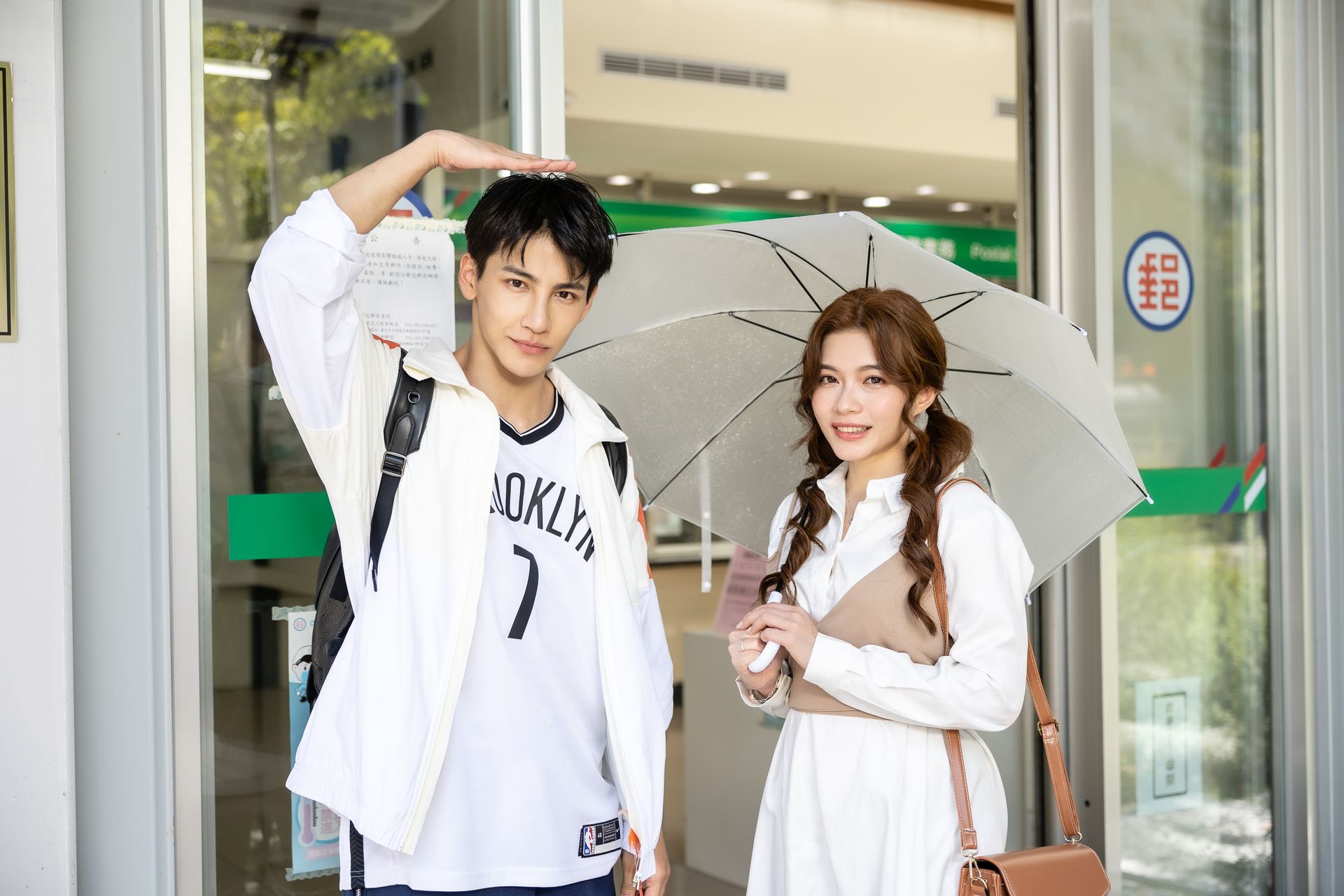 Man measures height with hand above woman holding an umbrella outside. Building entrance in background.
