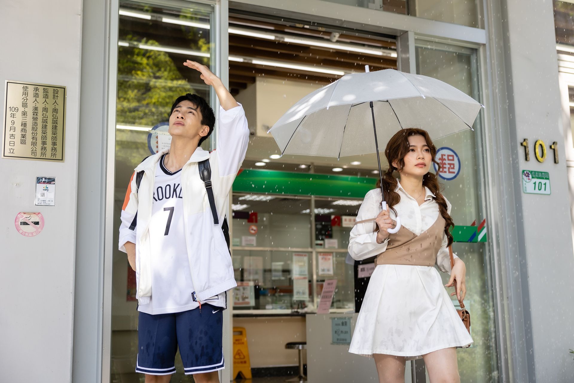 Man reaching up, woman holding umbrella in front of a convenience store.