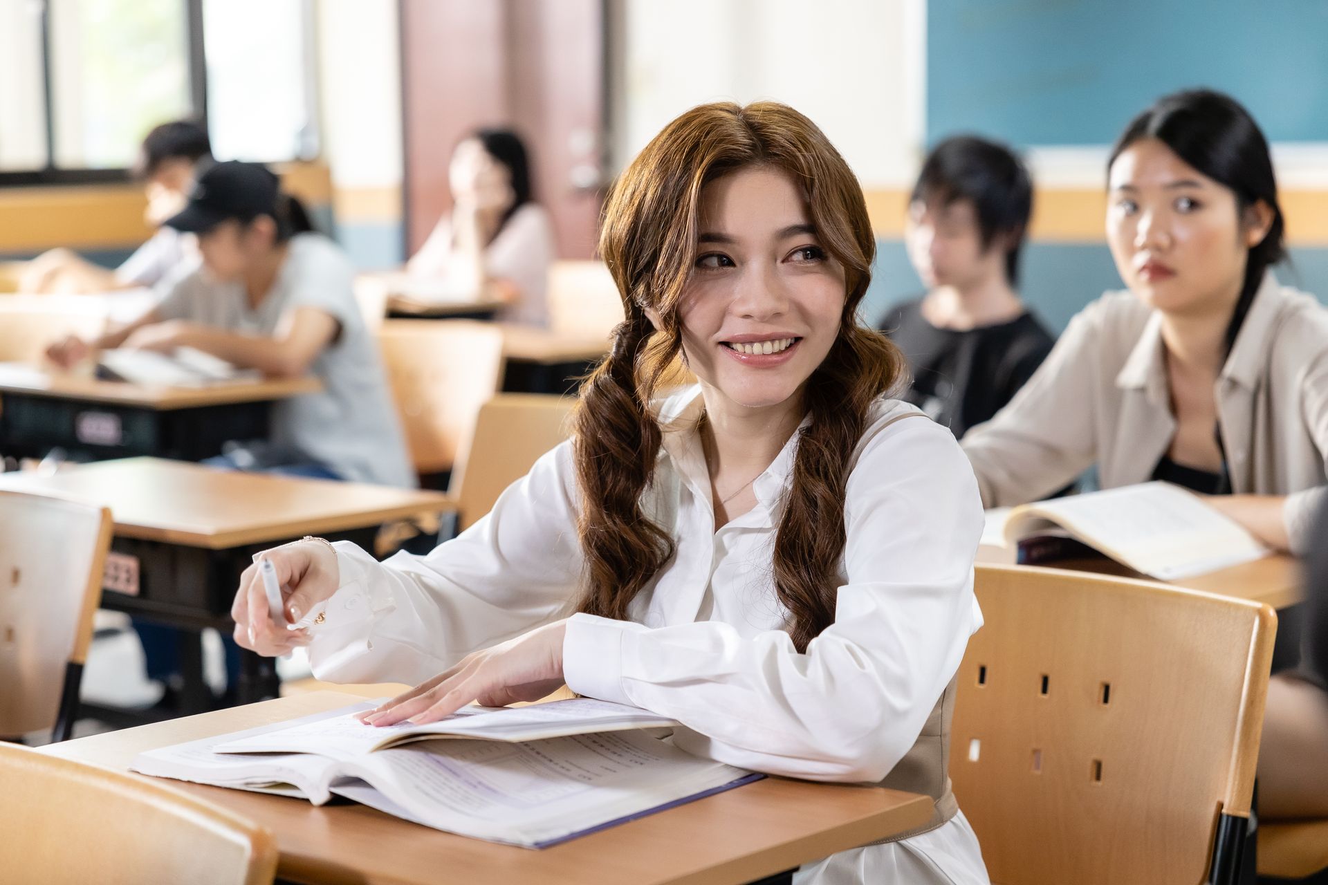 Woman with pigtails smiles in classroom, looking at papers. Students in background.