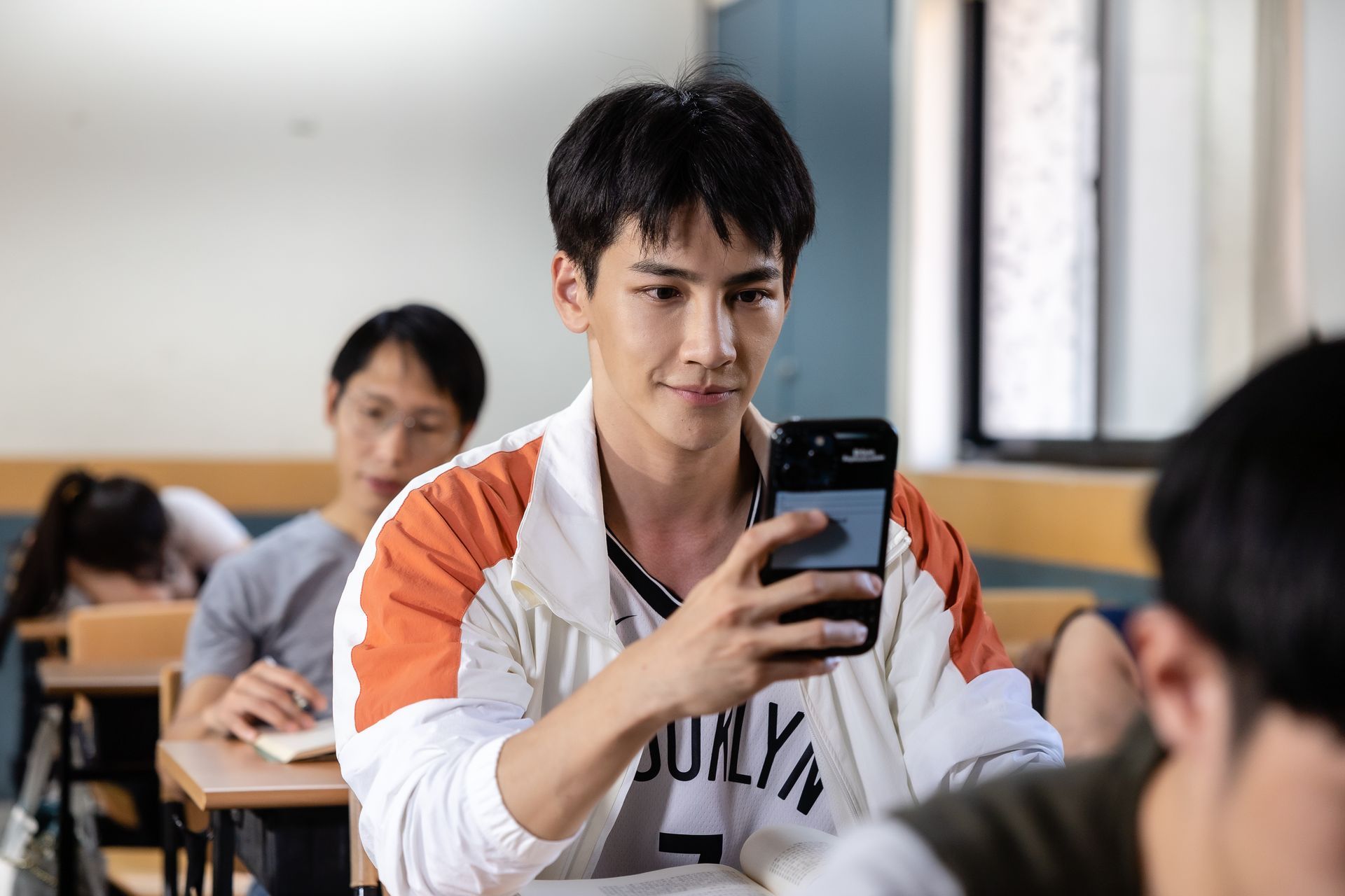 A student in a classroom looks at a smartphone; others in the background are studying or resting.