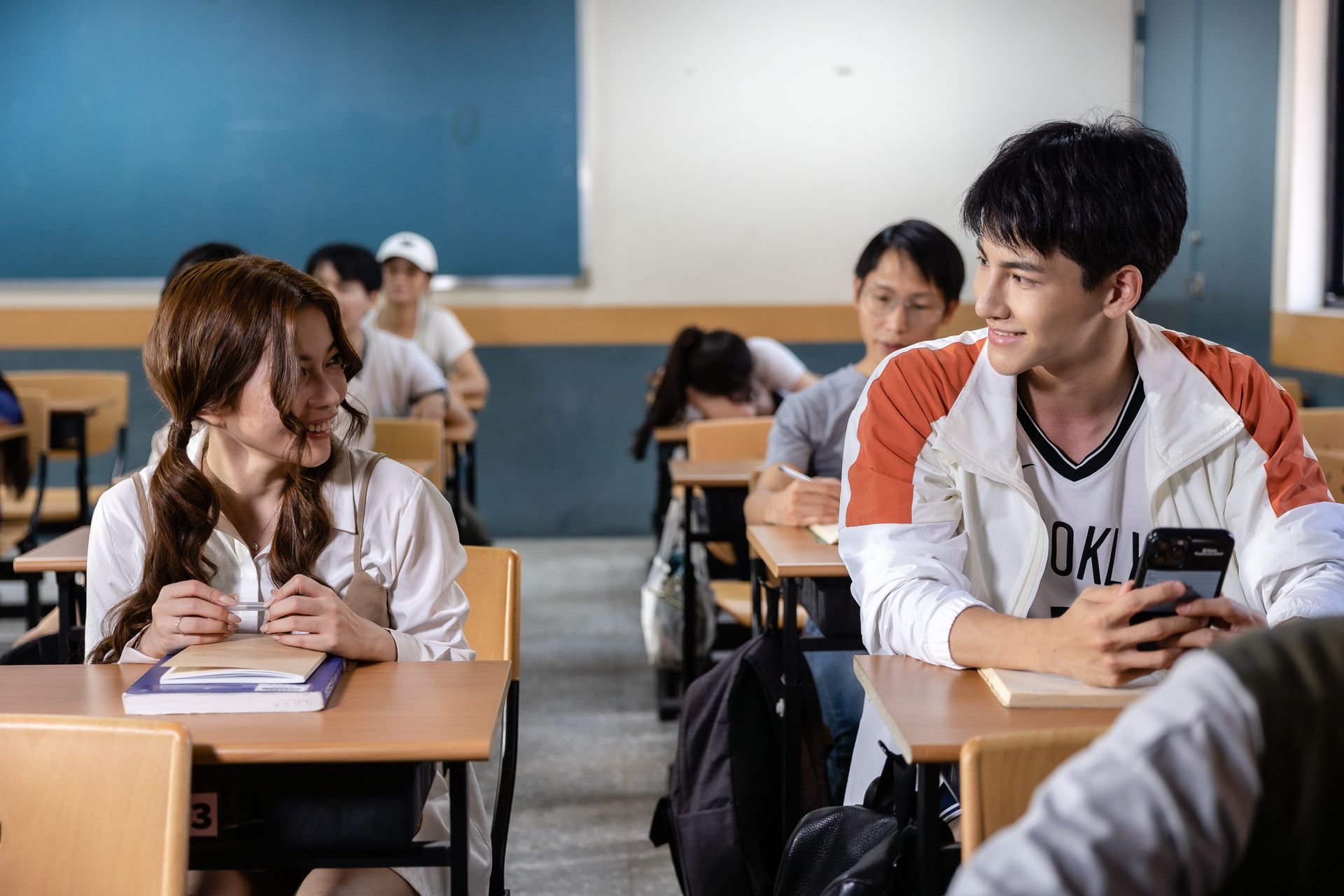 Two students in a classroom look at each other and smile. The male holds a phone. Others study.