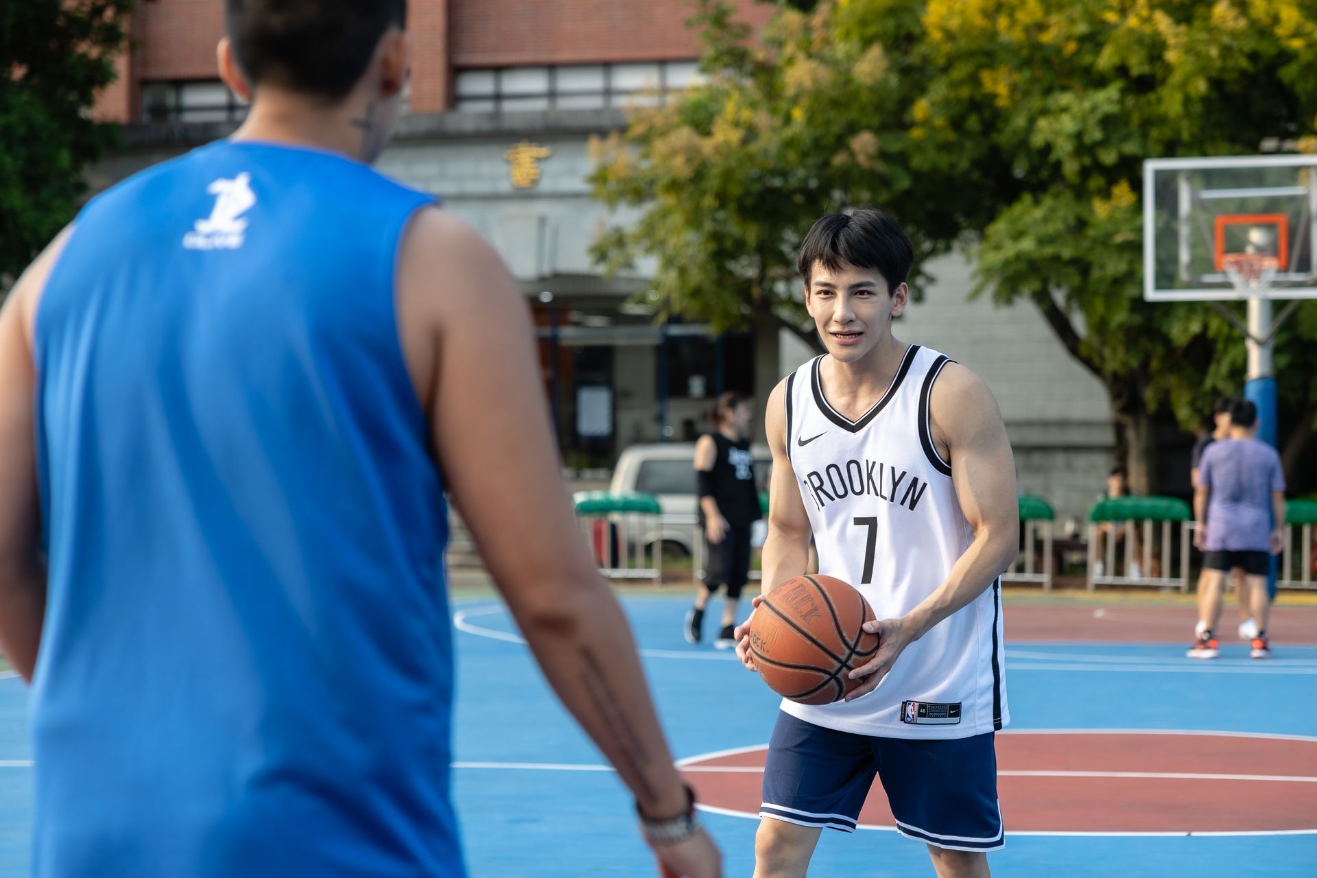 Two men playing basketball outdoors. One in blue, the other in a Brooklyn Nets jersey, holding the ball, looking towards the other.