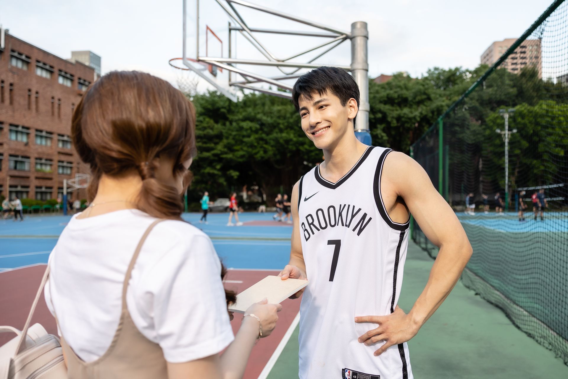 Man in Brooklyn basketball jersey smiles, holding paper, speaking to a person at a basketball court.