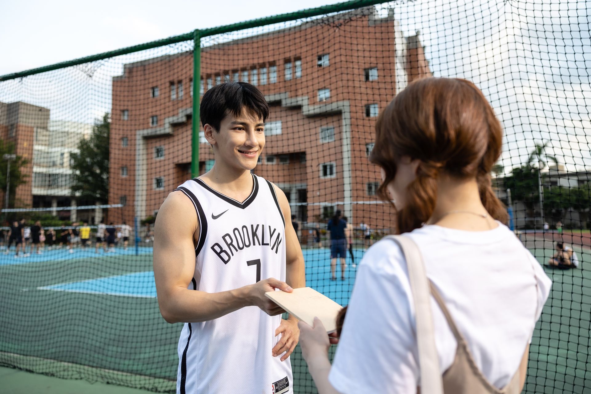 Basketball player in Brooklyn jersey smiling, accepting a paper from a person on a court. Brick building in background.