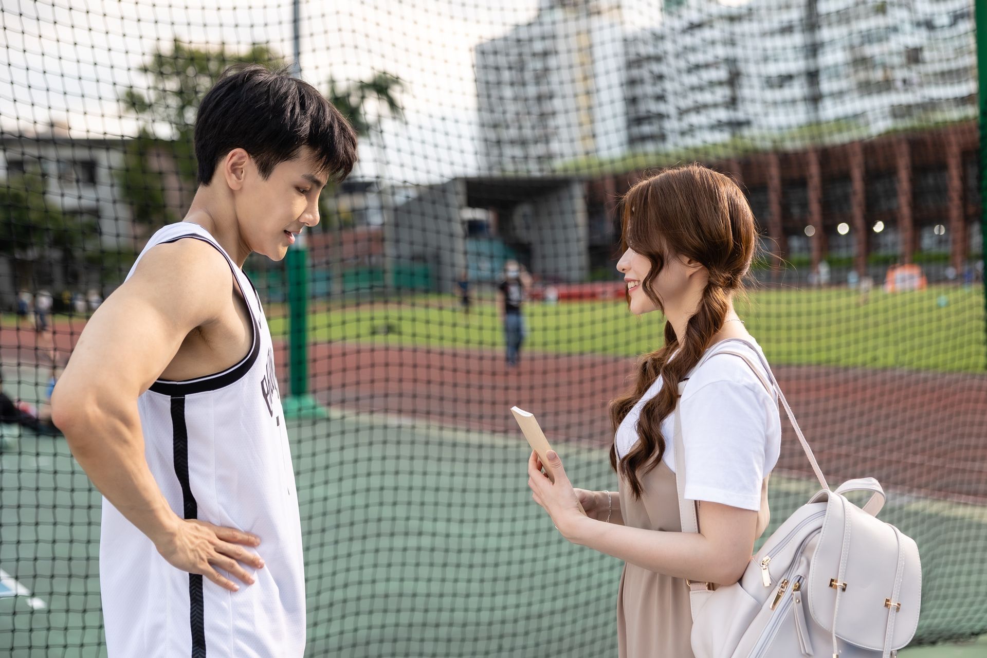 Man in basketball jersey and woman with a smartphone on a sports field, talking.