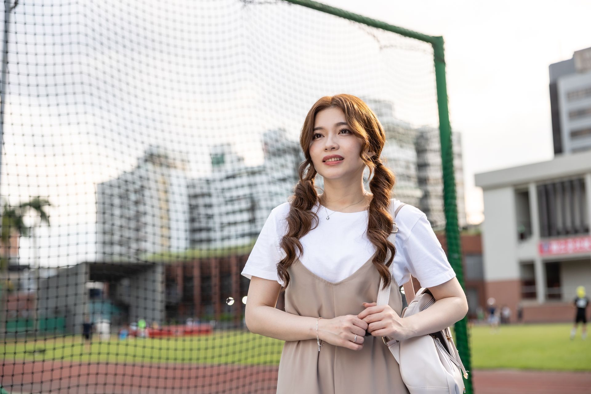 Woman with pigtails stands on a field, wearing a white shirt and overalls, bag over shoulder.