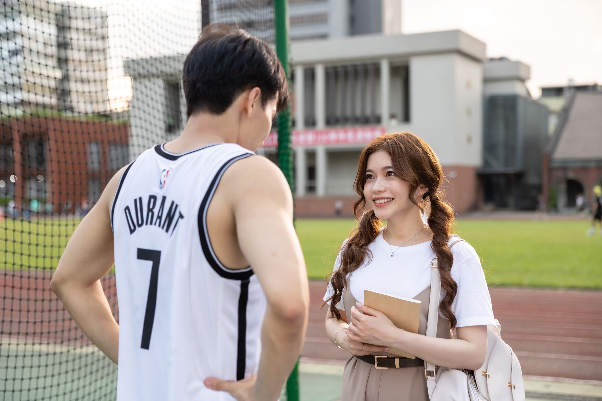 A man in a basketball jersey and a woman with pigtails converse on a school athletic field.
