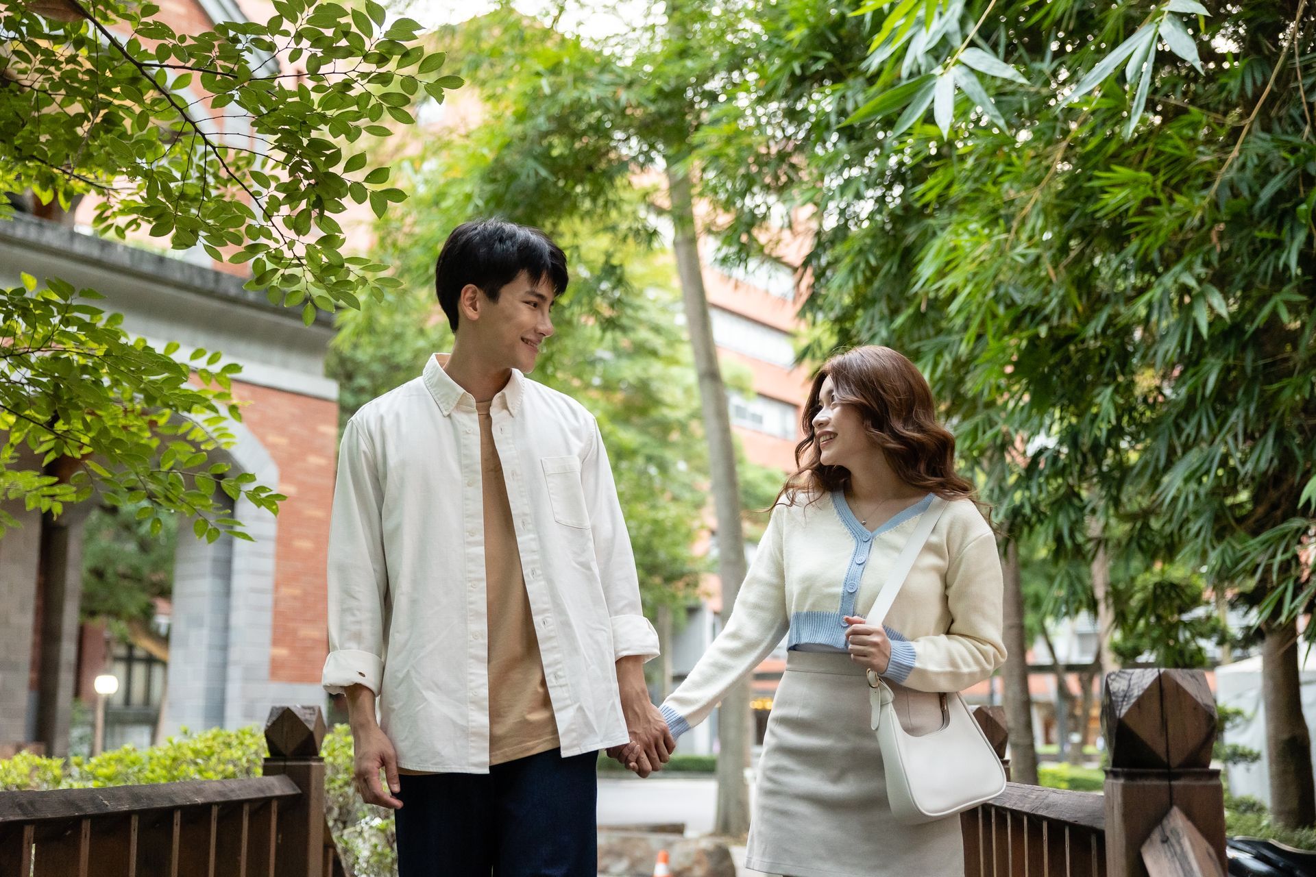Couple holding hands, walking on a bridge, smiling. Green trees and building in the background. Sunny day.