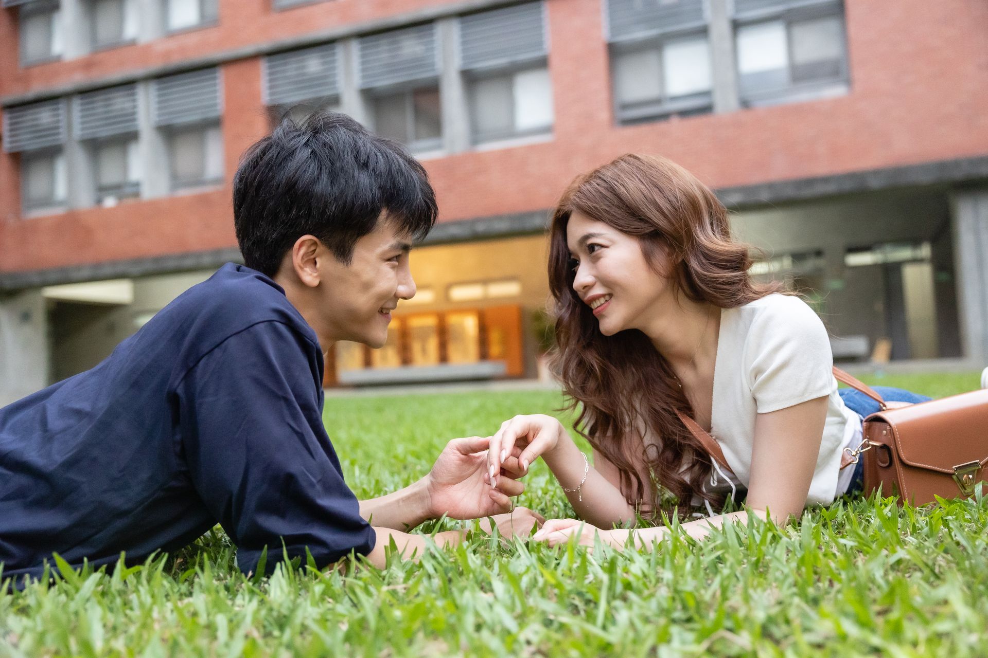 Couple lying on grass, holding hands, smiling at each other in front of a brick building.