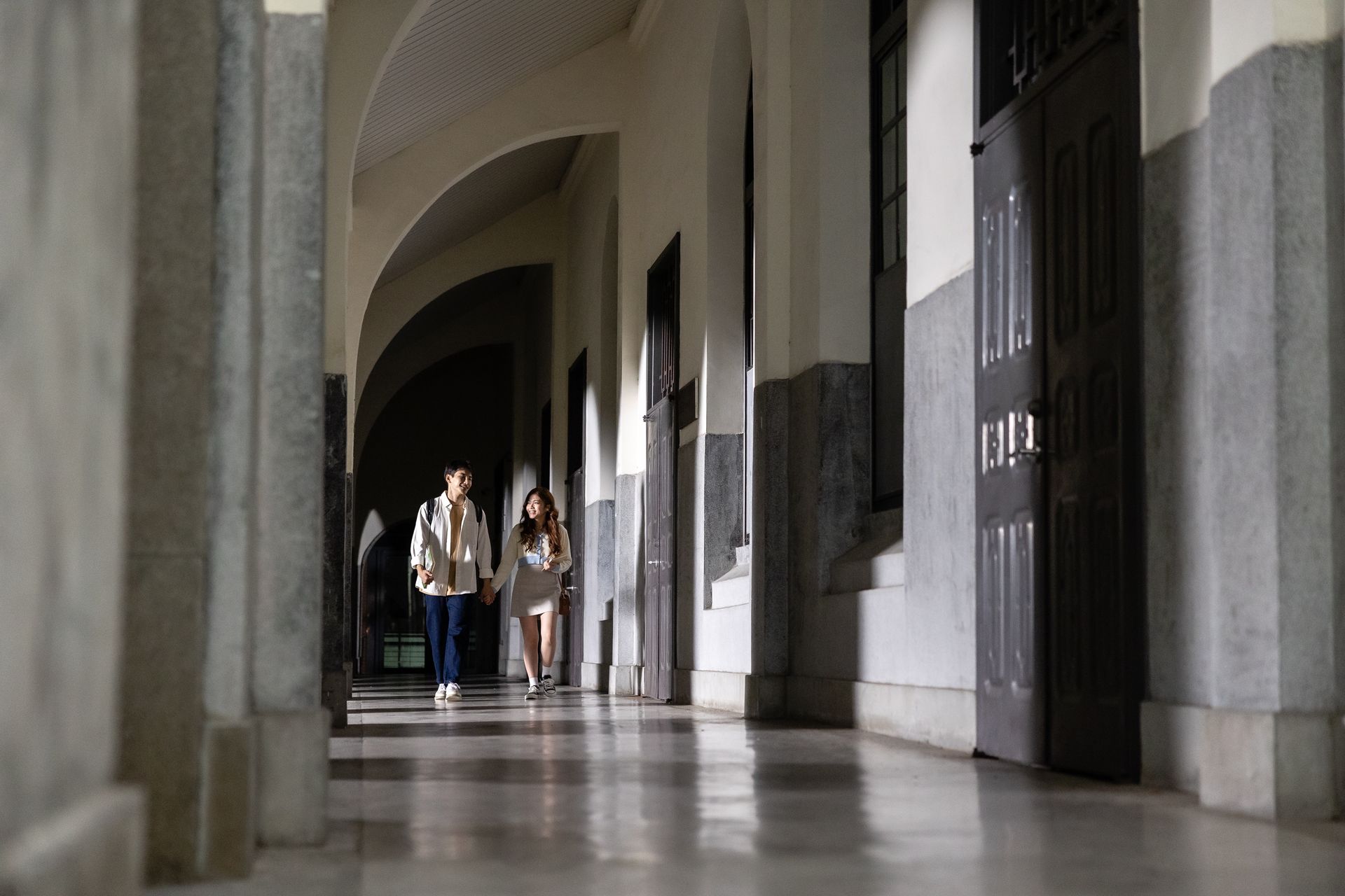Two people walk down a long hallway with arched ceilings and doors.