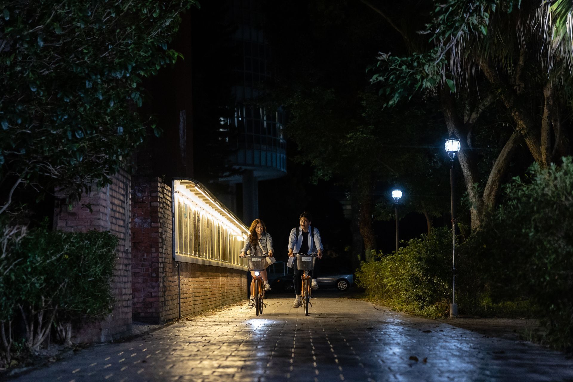 Two people biking on a dark path lit by streetlights. Brick wall and trees flank the path.