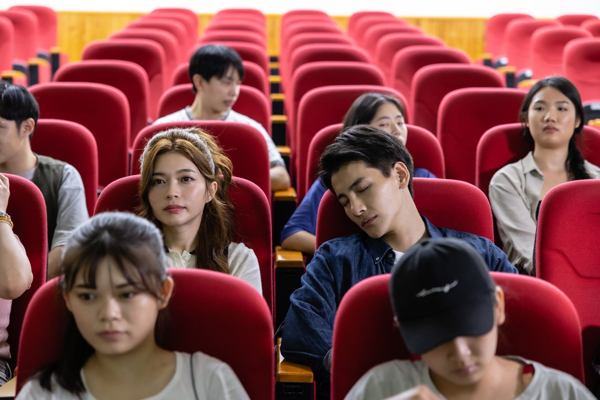 Auditorium with rows of red seats. Students sit, some attentive, others sleeping, in a lecture hall.