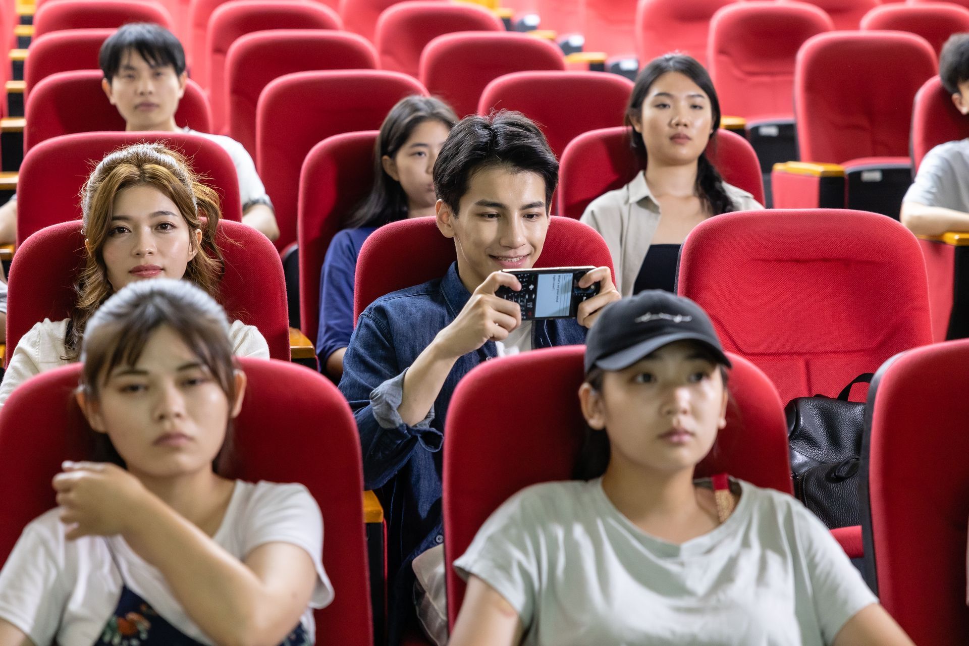 People in red theater seats, one taking a picture with a phone.