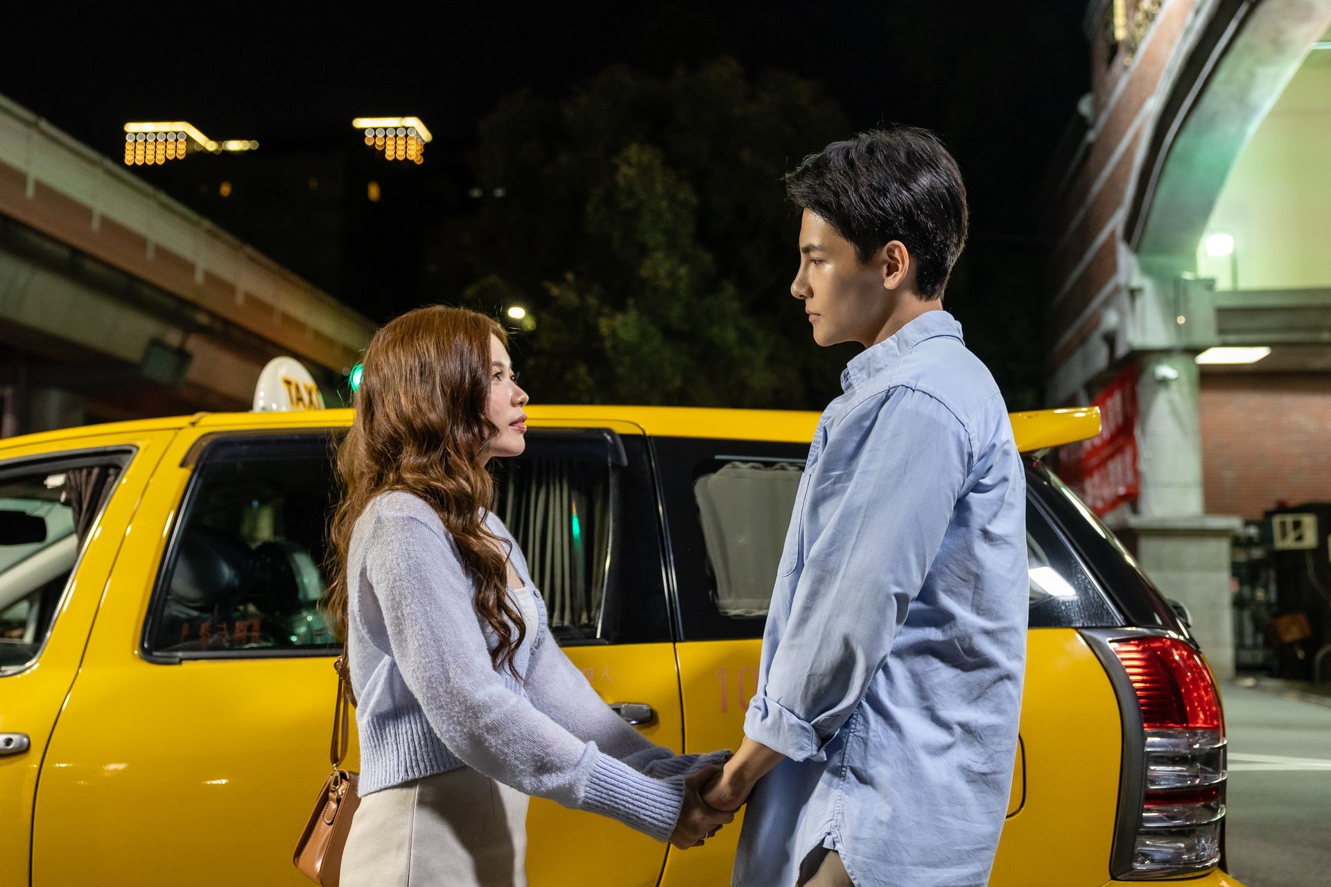 Couple holding hands near a yellow taxi at night. Buildings and streetlights visible.