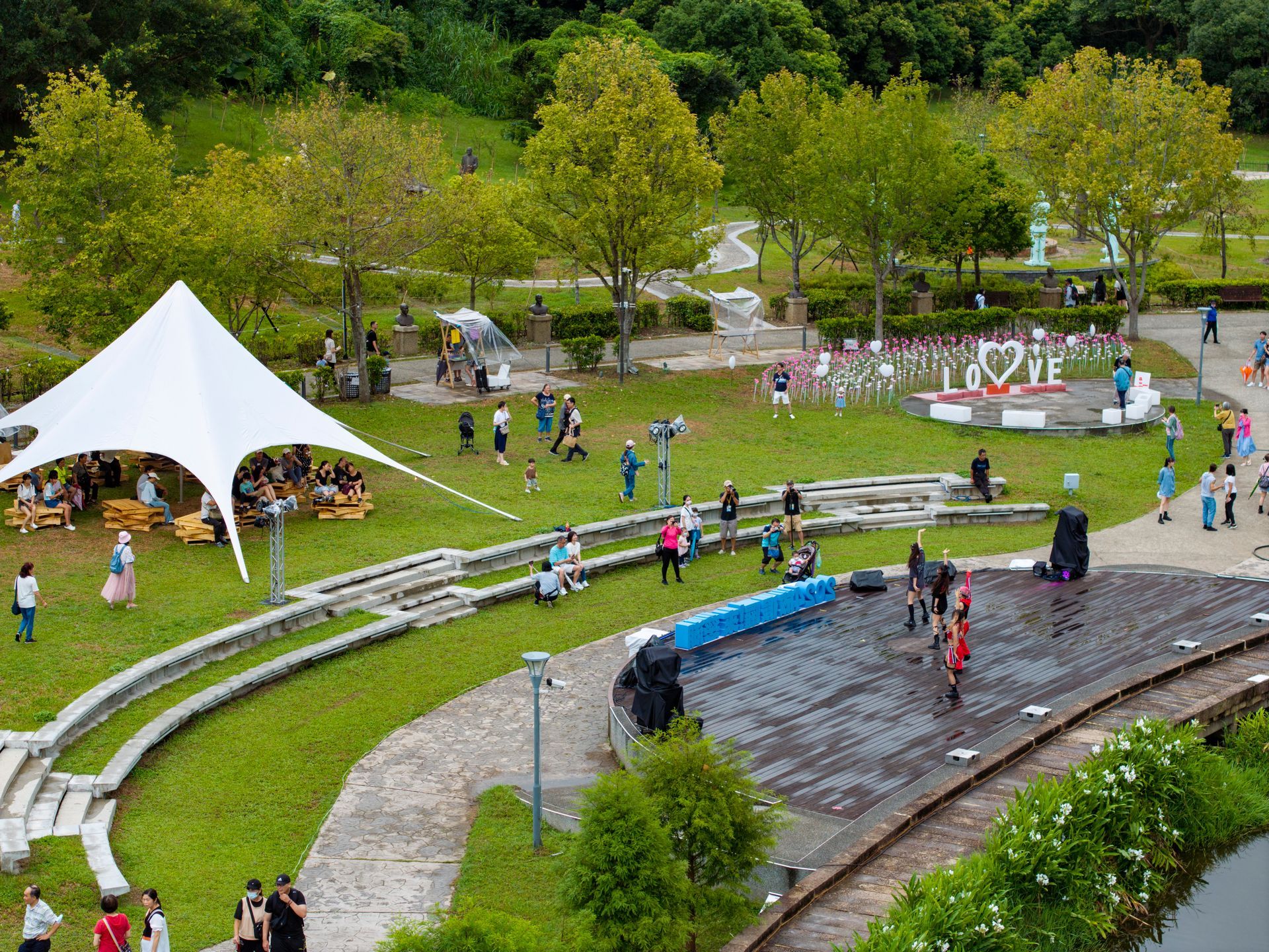 Park with people, stage, tent, and landscaping. White tent on grass, people walking, trees, and
