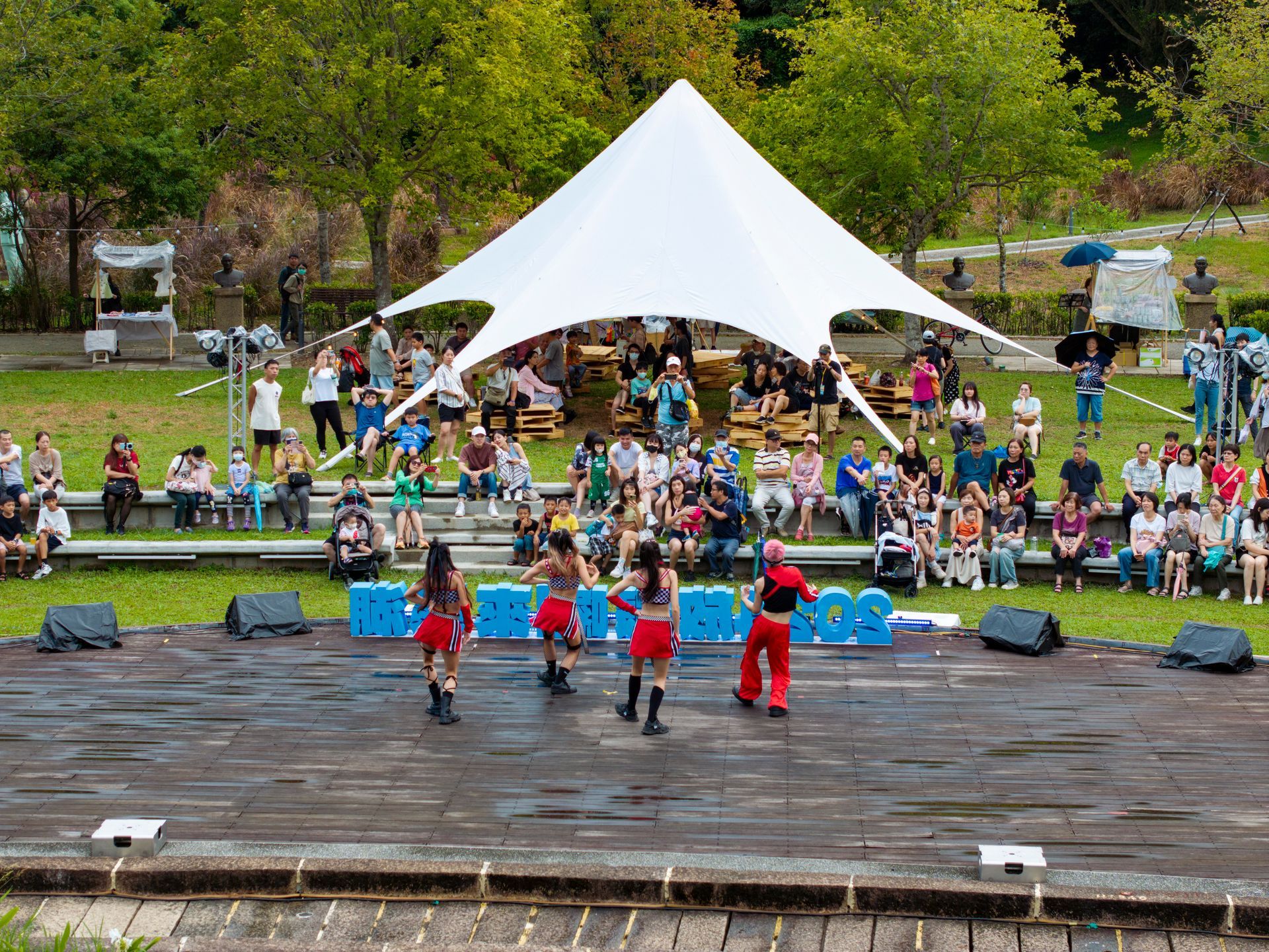 Dancers perform on a stage with a tent overhead, an audience watches in an outdoor amphitheater.