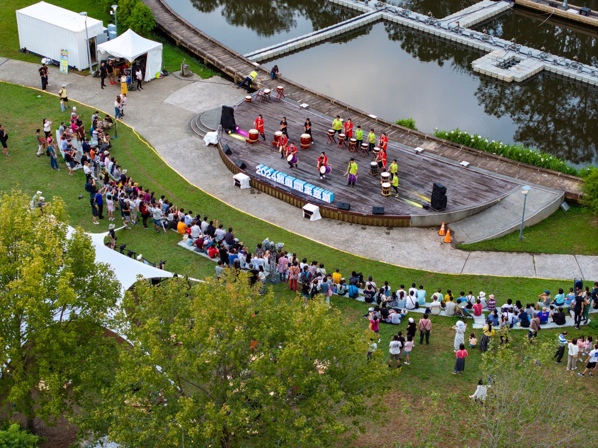 Outdoor performance: Musicians on stage; audience on lawn; water in background.