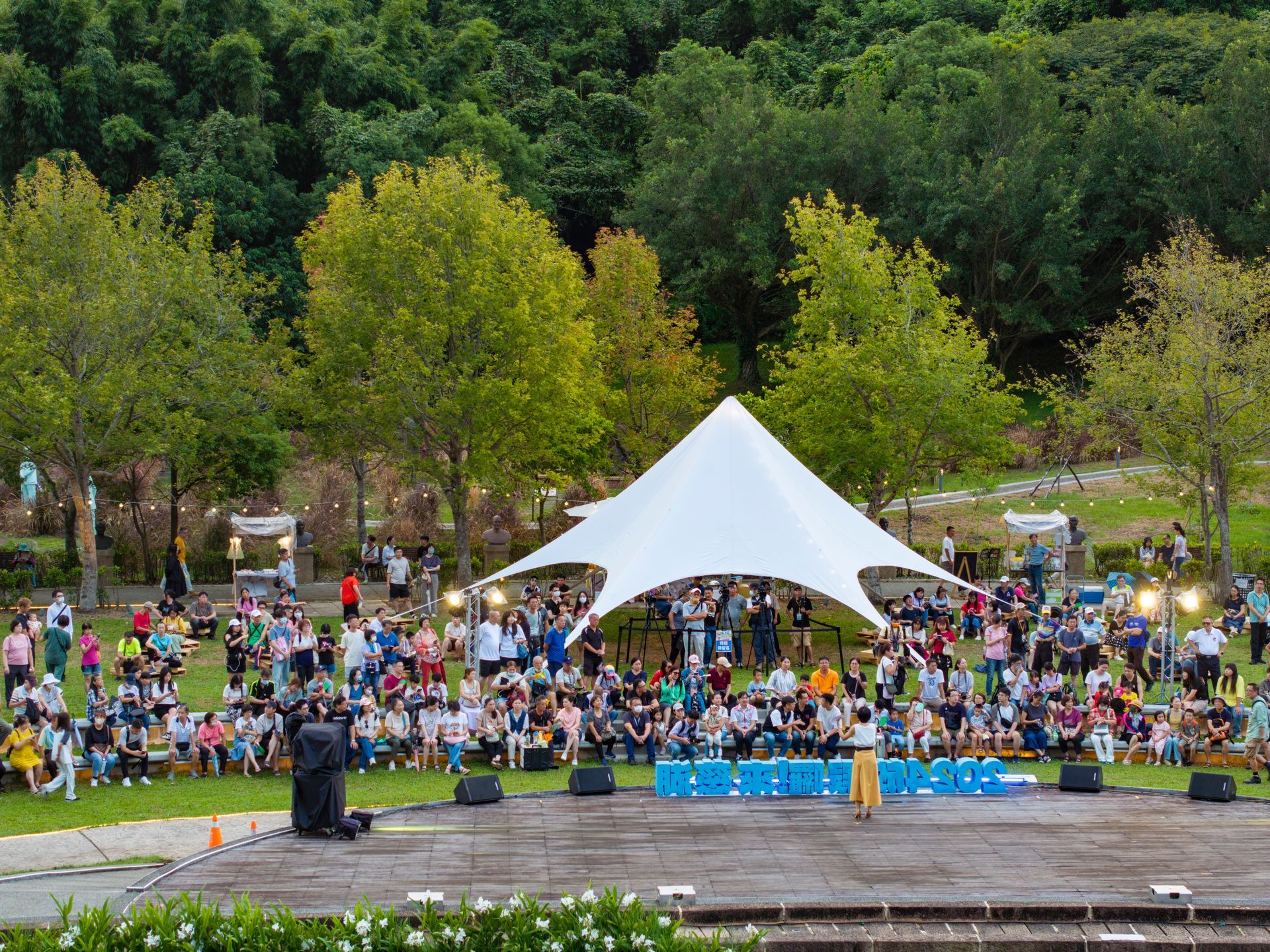 Outdoor event with a white tent, stage, and crowd on a grassy hill, surrounded by trees.