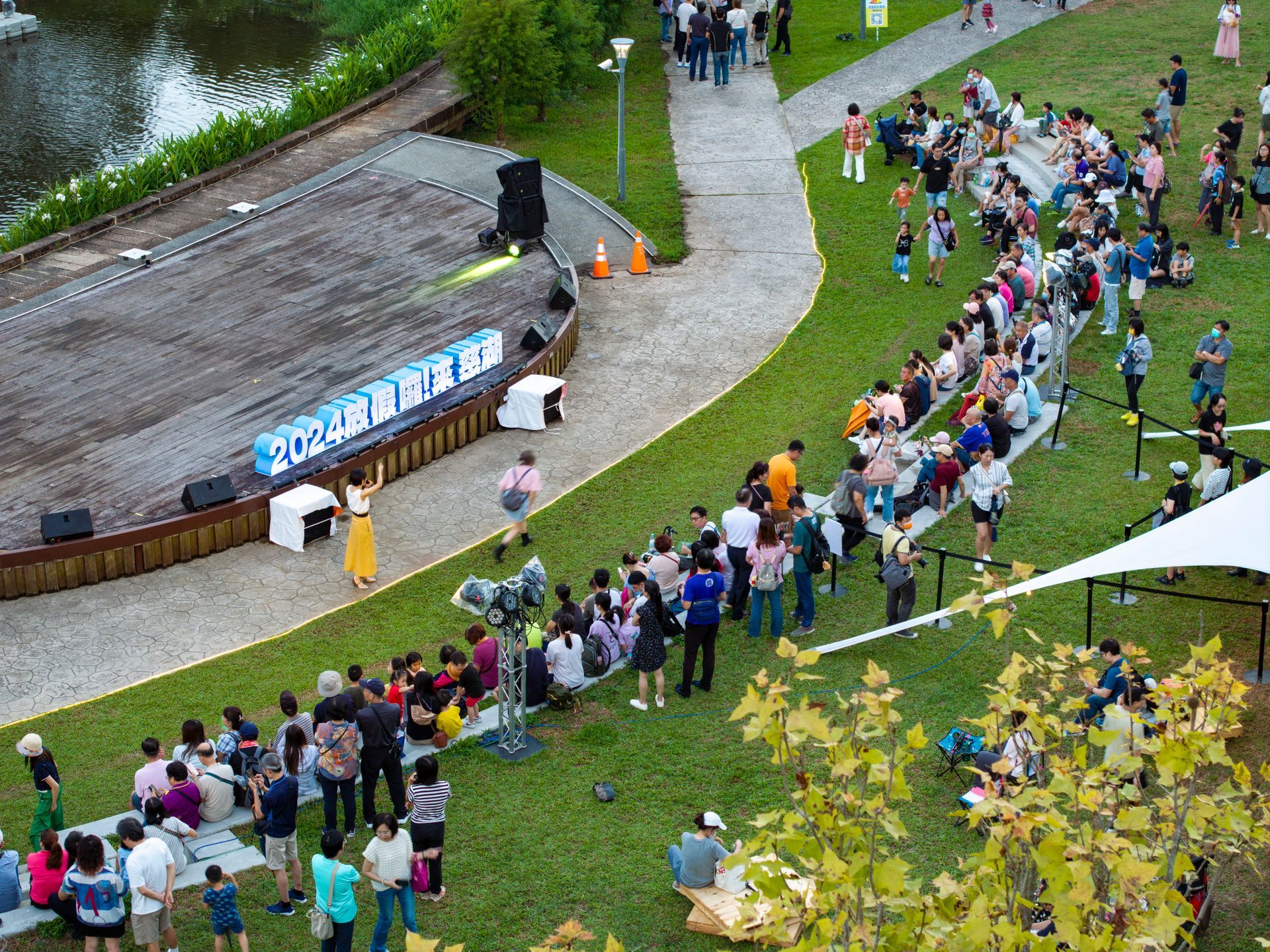 Outdoor event with crowd, stage, and path near water, greenery. People sit/stand, some watch stage.