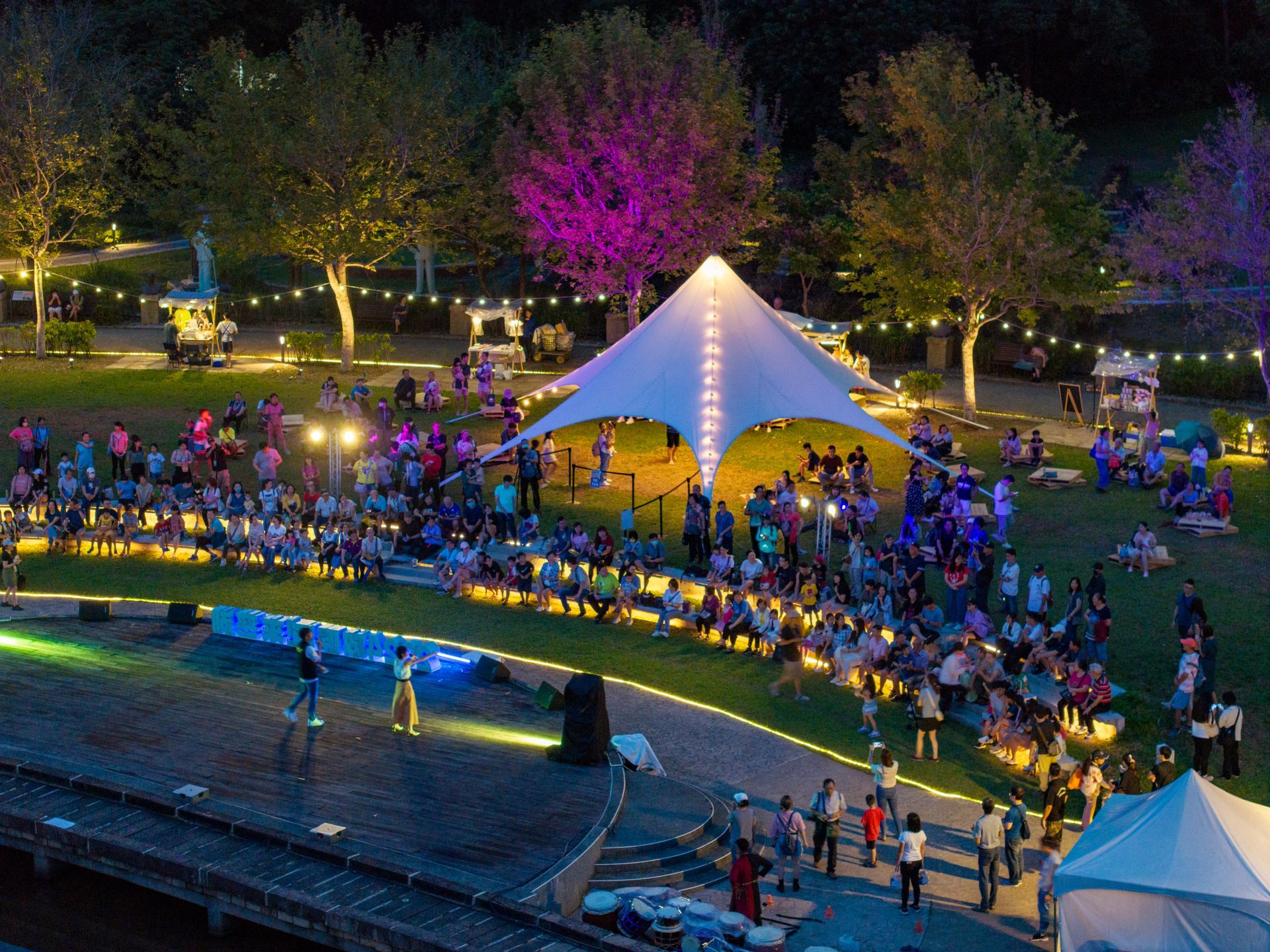 Outdoor evening event under a lit tent. People seated on a grassy hillside, stage with performers.