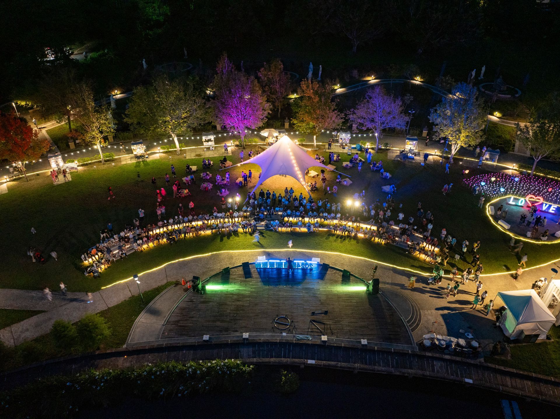 Nighttime aerial view of an outdoor event. People gathered around a stage, illuminated trees, and food vendors.