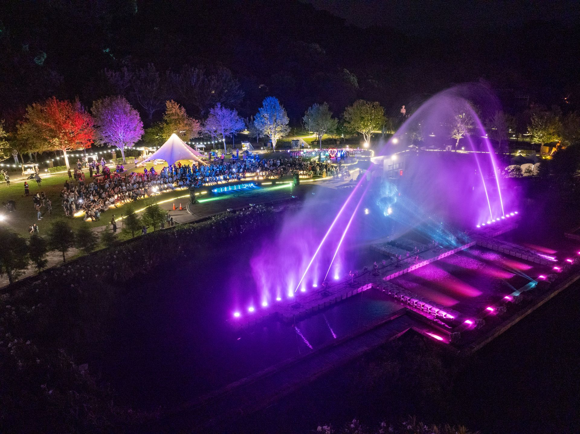 A nighttime fountain show with colorful lights, surrounded by trees and an audience.