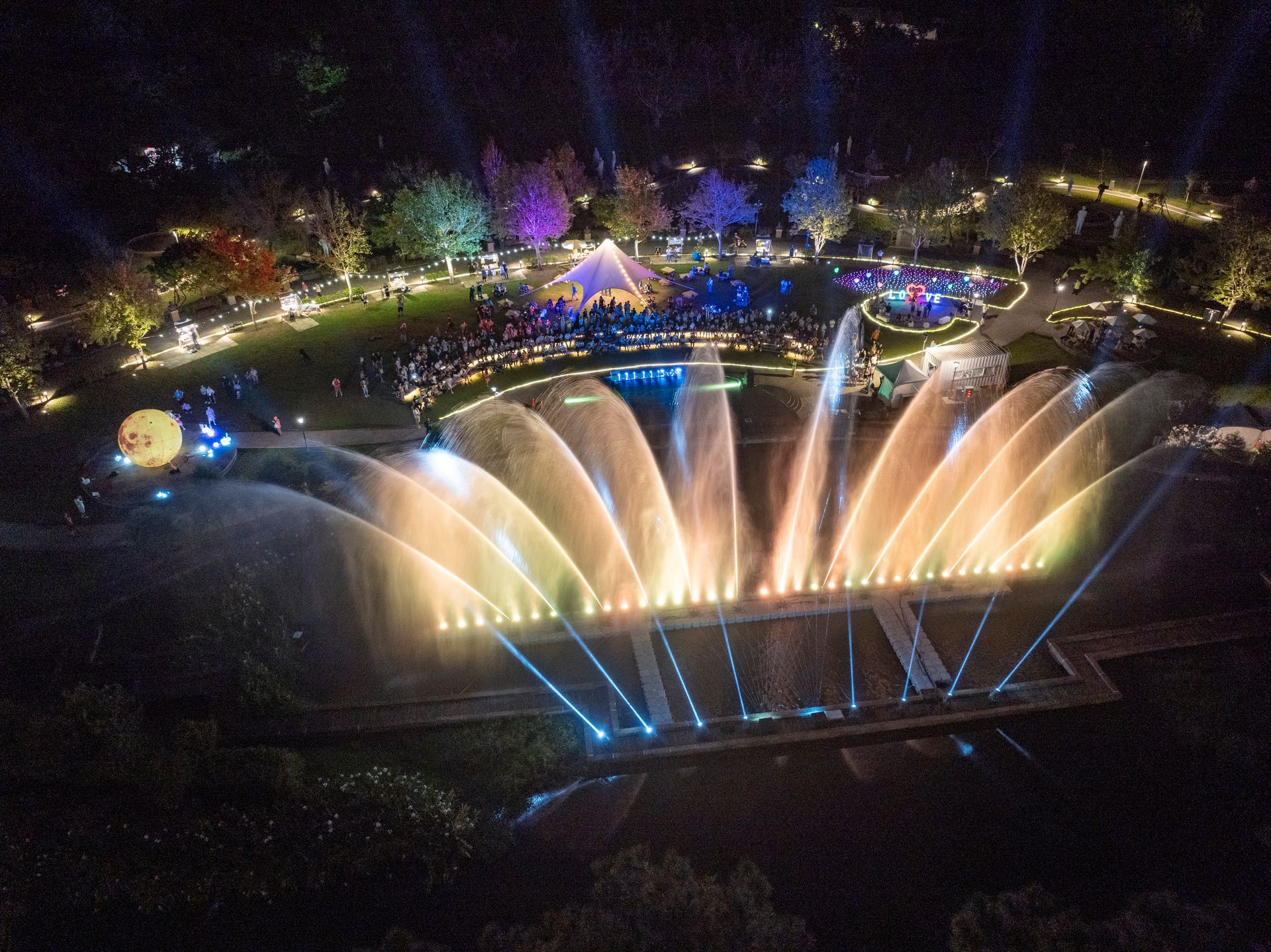 Nighttime view of a water fountain show with colorful lights, trees, and a crowd.