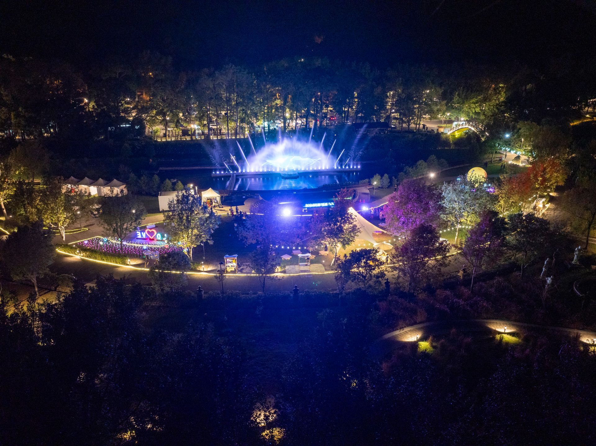 Night aerial view of a park with a lit stage, water fountain, and colorful lights.