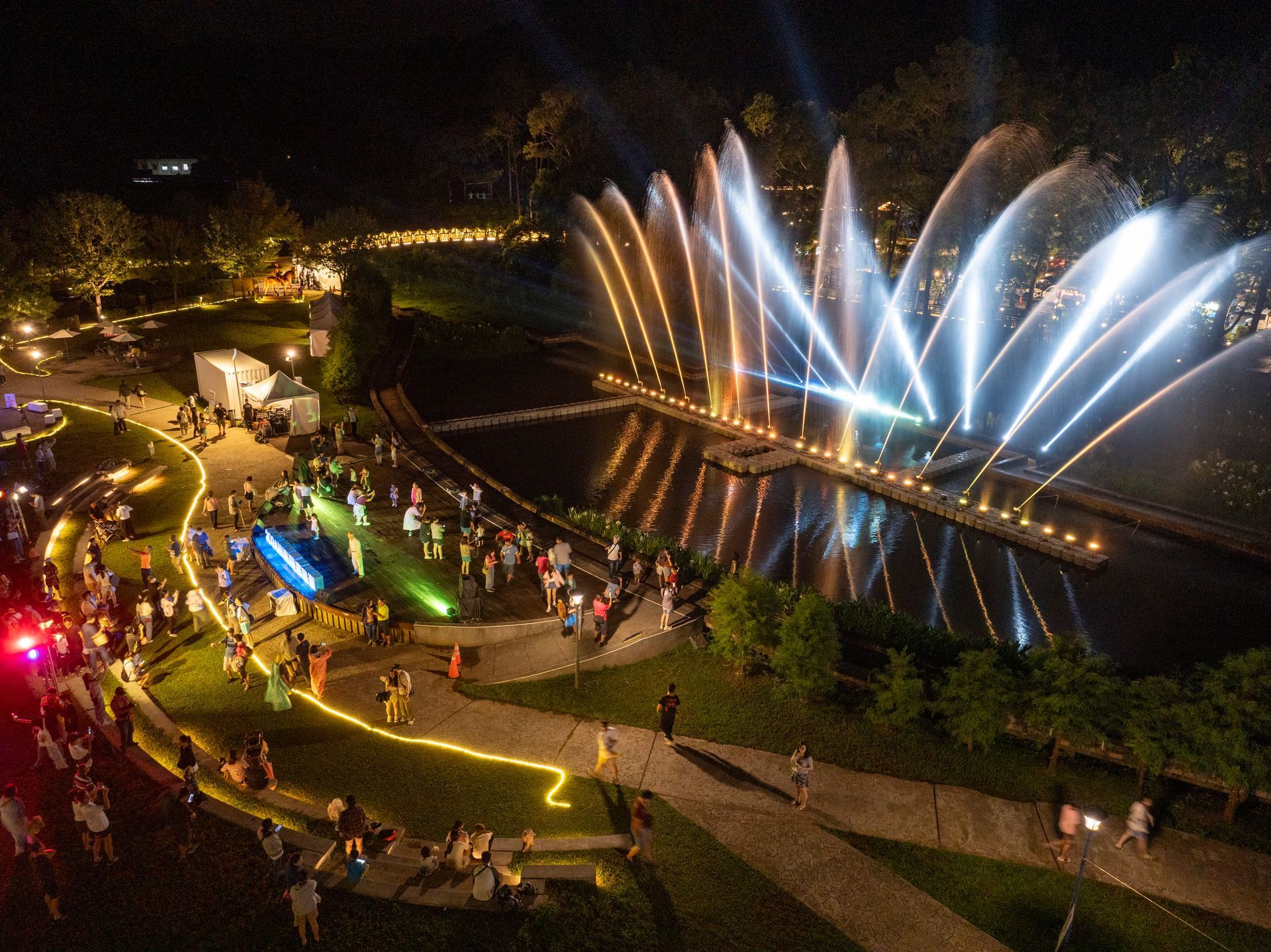 Night view of a water fountain show with illuminated jets of water, lights, and people on a lawn.