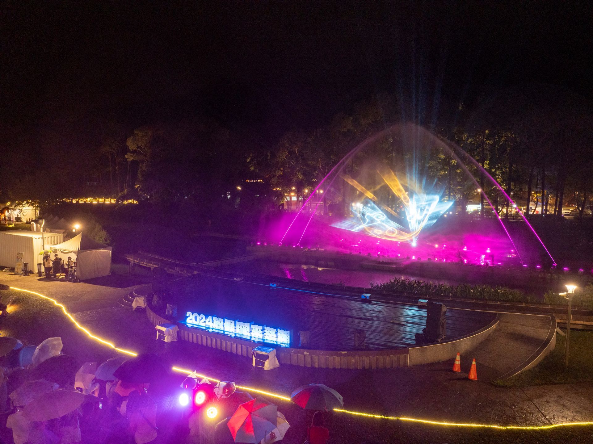 Night view of a fountain with colorful lights. People with umbrellas watch the show.