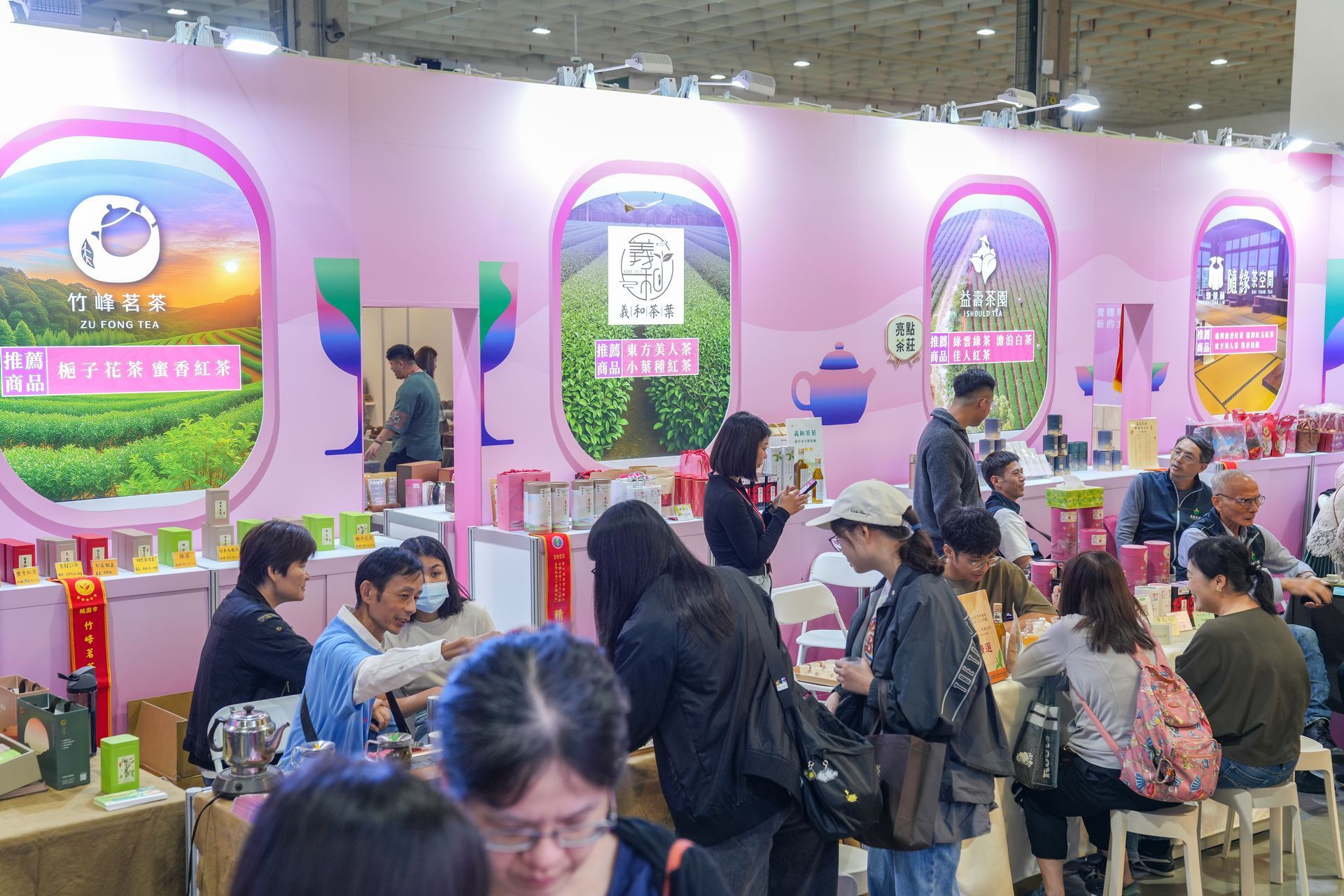 People browsing a tea exhibition booth with pink accents, tea product displays, and staff assisting customers.