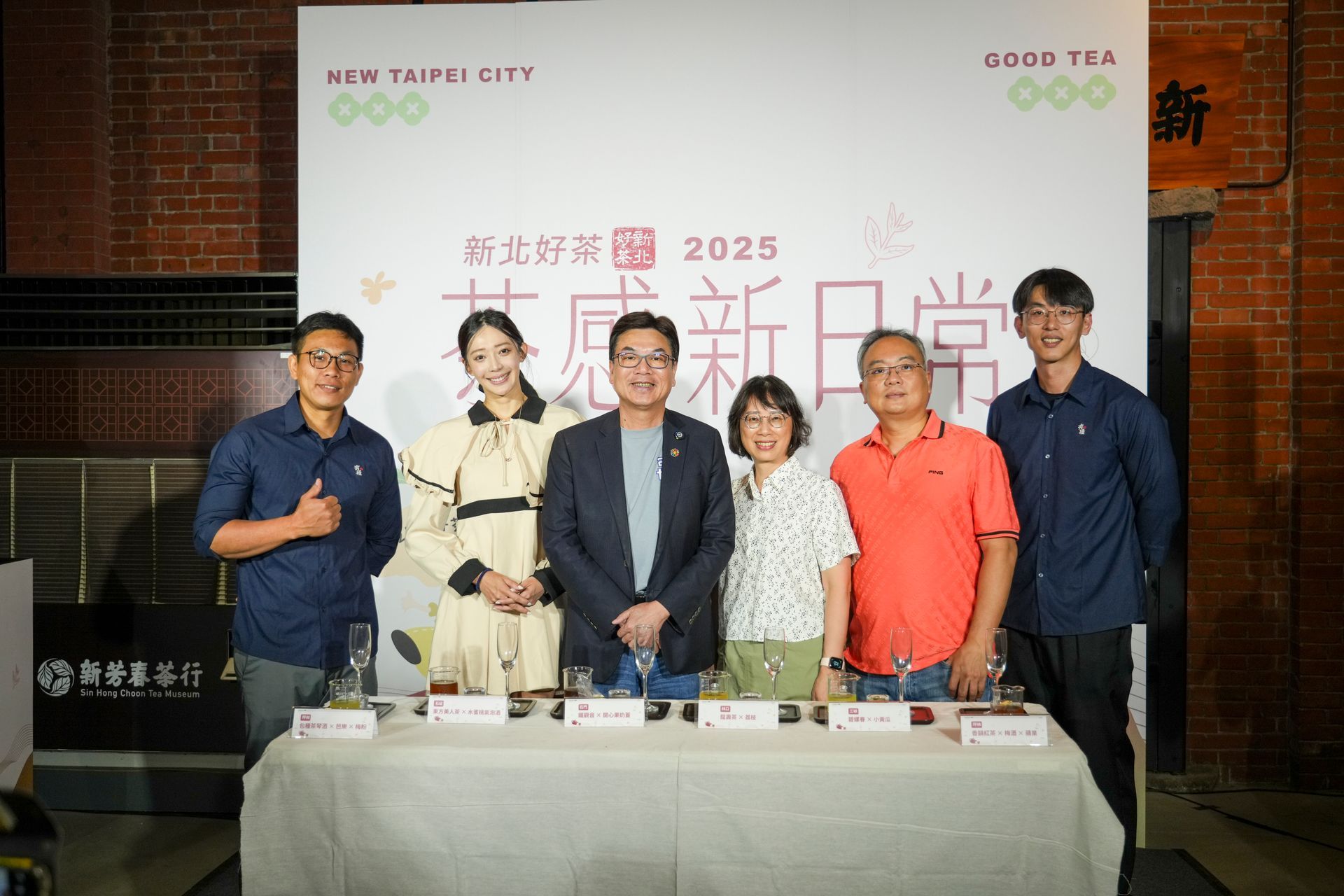 Group of people at a table, with a banner behind them. They are smiling, with microphones and tea cups.